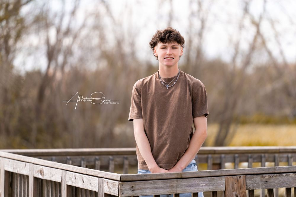 Someone in a brown t-shirt stands on a wooden boardwalk against a blurred natural backdrop on a sunny day.
