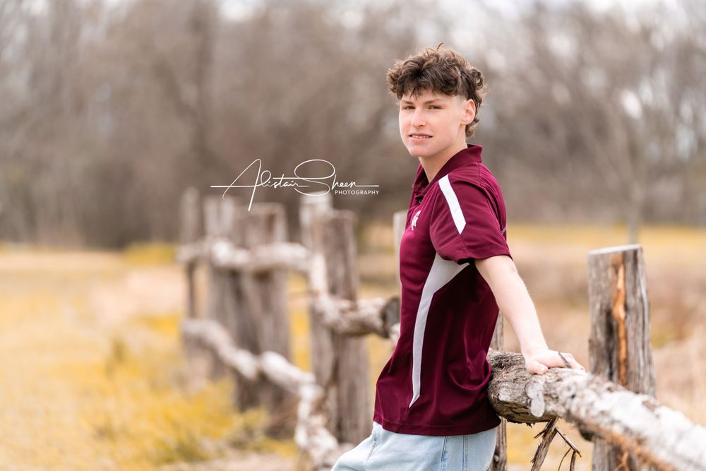 Young person in a burgundy shirt leans against a rustic wooden fence in an autumn field with blurred trees in background.