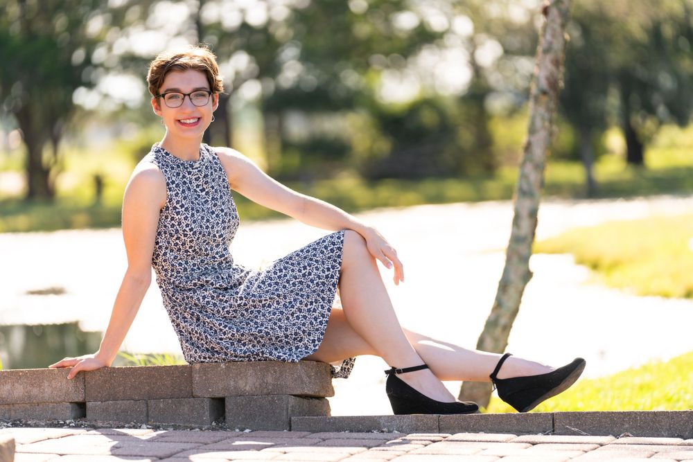 A casual outdoor portrait sitting on wood beams in a patterned dress and flats.
