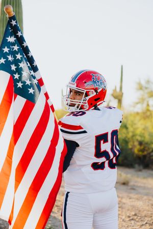Football player in white jersey celebrates with American flag among desert cacti.