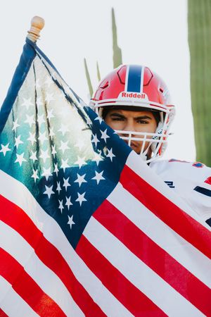 Football player in red helmet holds American flag against green background.