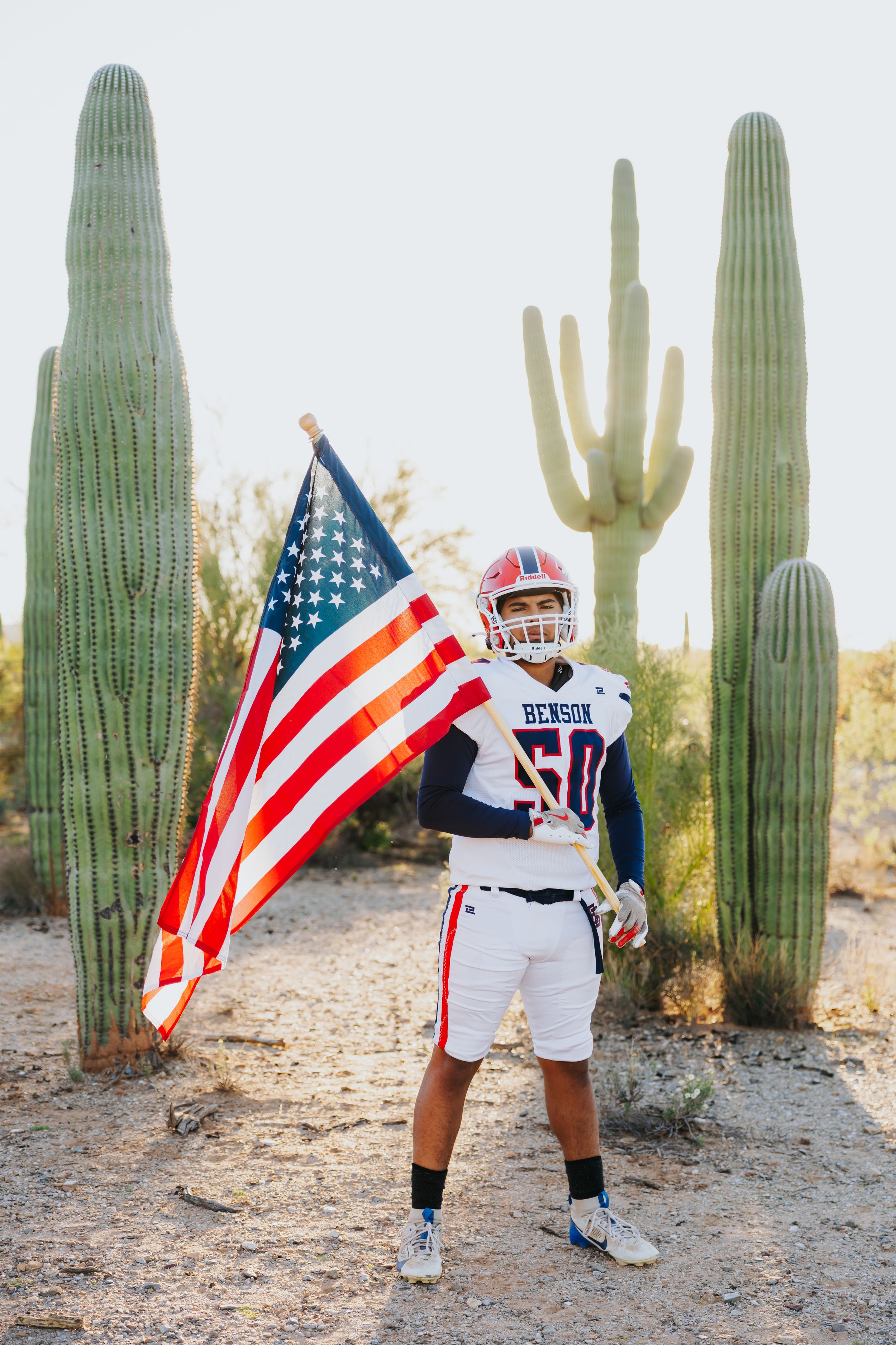 Football player in white uniform waves American flag among desert cacti landscape.