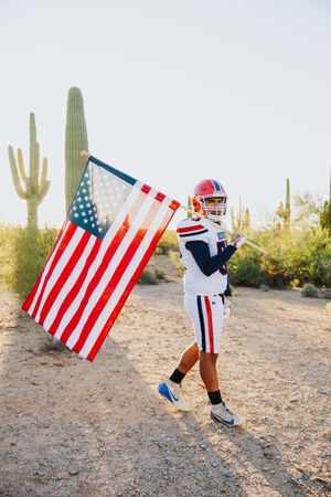 Football player carries American flag through desert landscape with saguaro cacti.