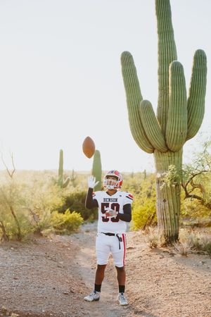 Football player throws football in desert setting surrounded by tall saguaro cacti.
