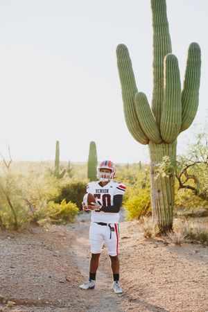 Football player in white uniform stands with football against desert landscape.