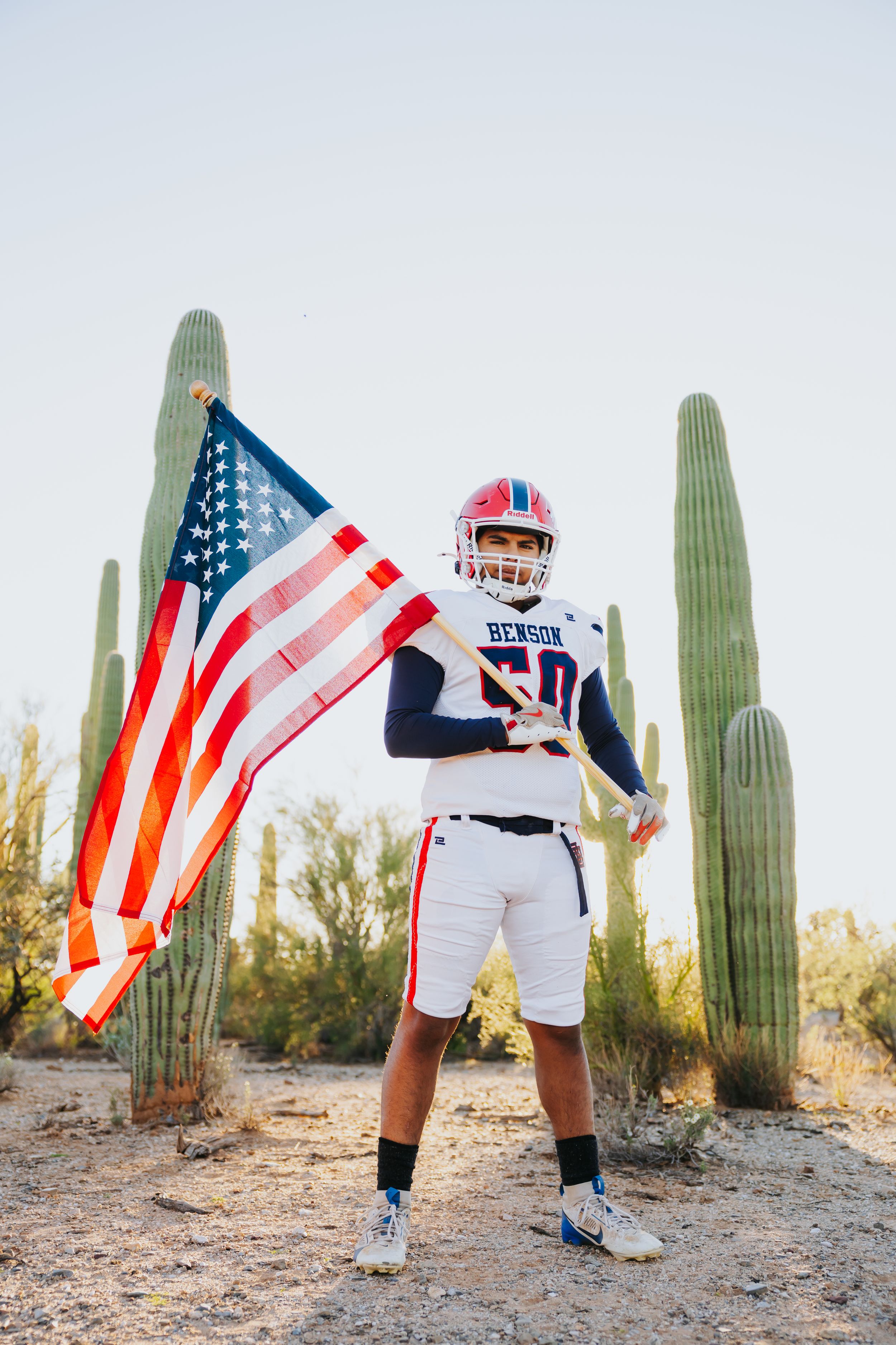 Athlete in white uniform holds American flag while posing between saguaro cacti.