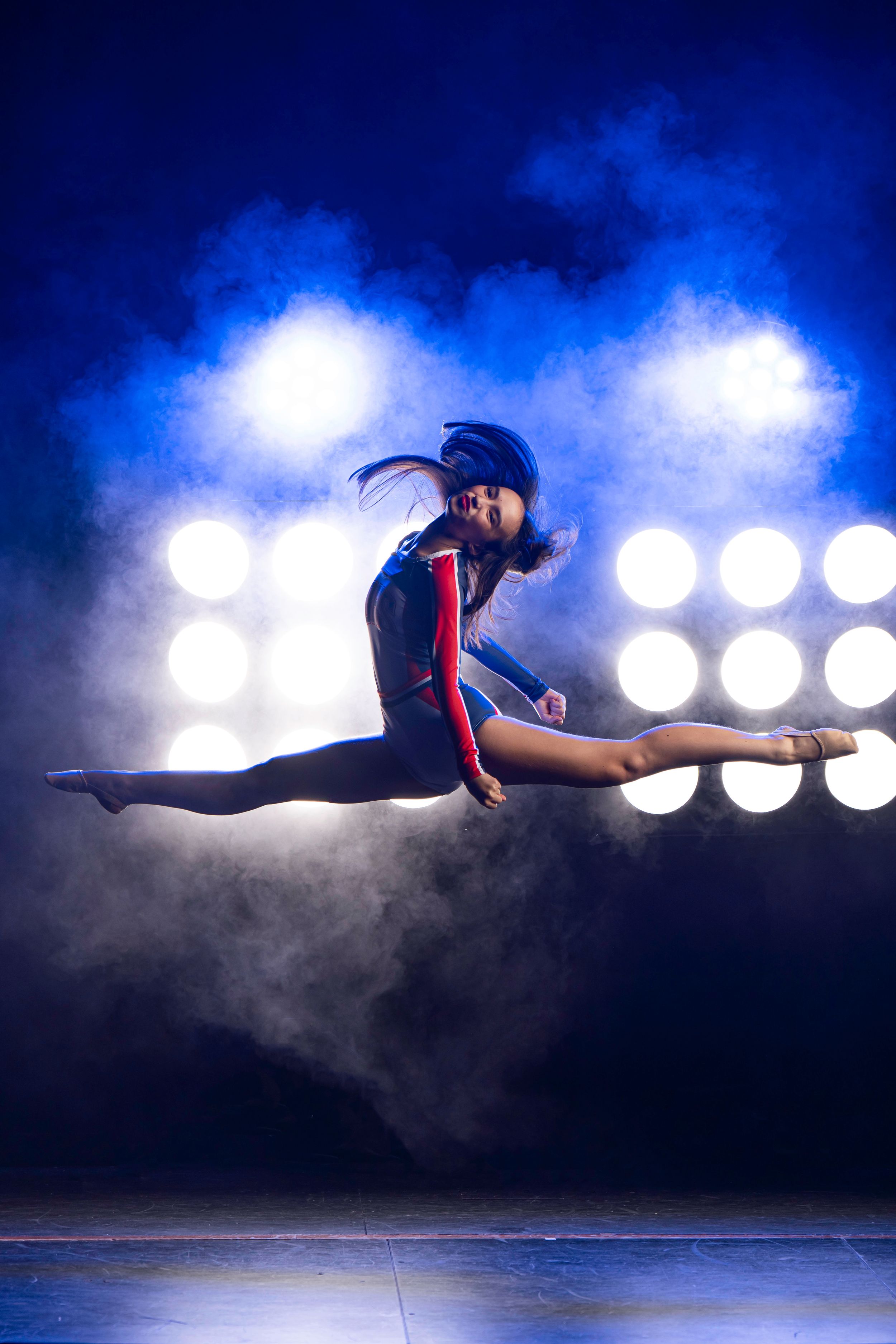 Athletic gymnast performs dramatic split jump in mid-air with spotlights and fog effects on dark stage.