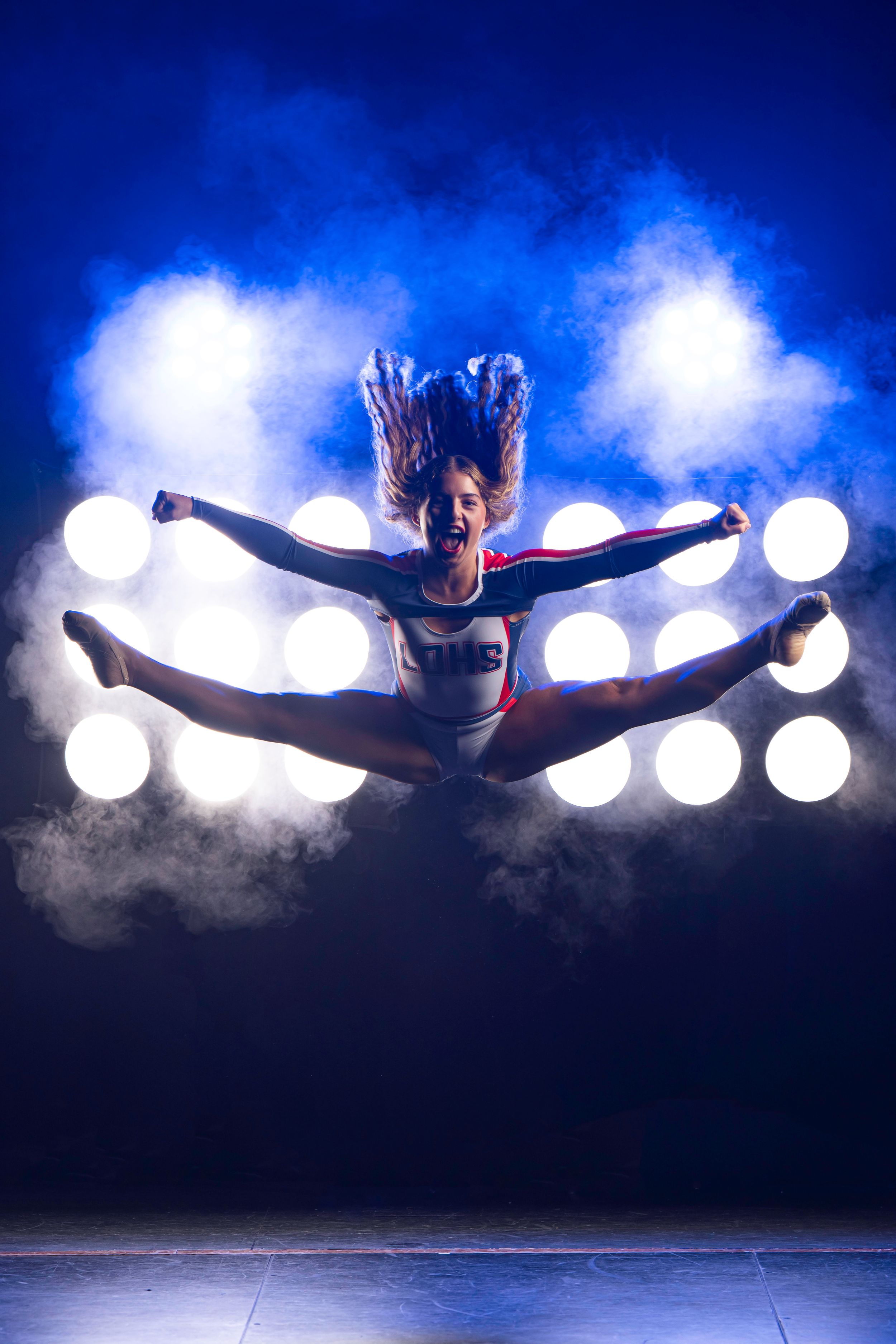 A high school dancer performs a powerful split leap against a dark blue background with glowing orbs of light and atmospheric fog effects.