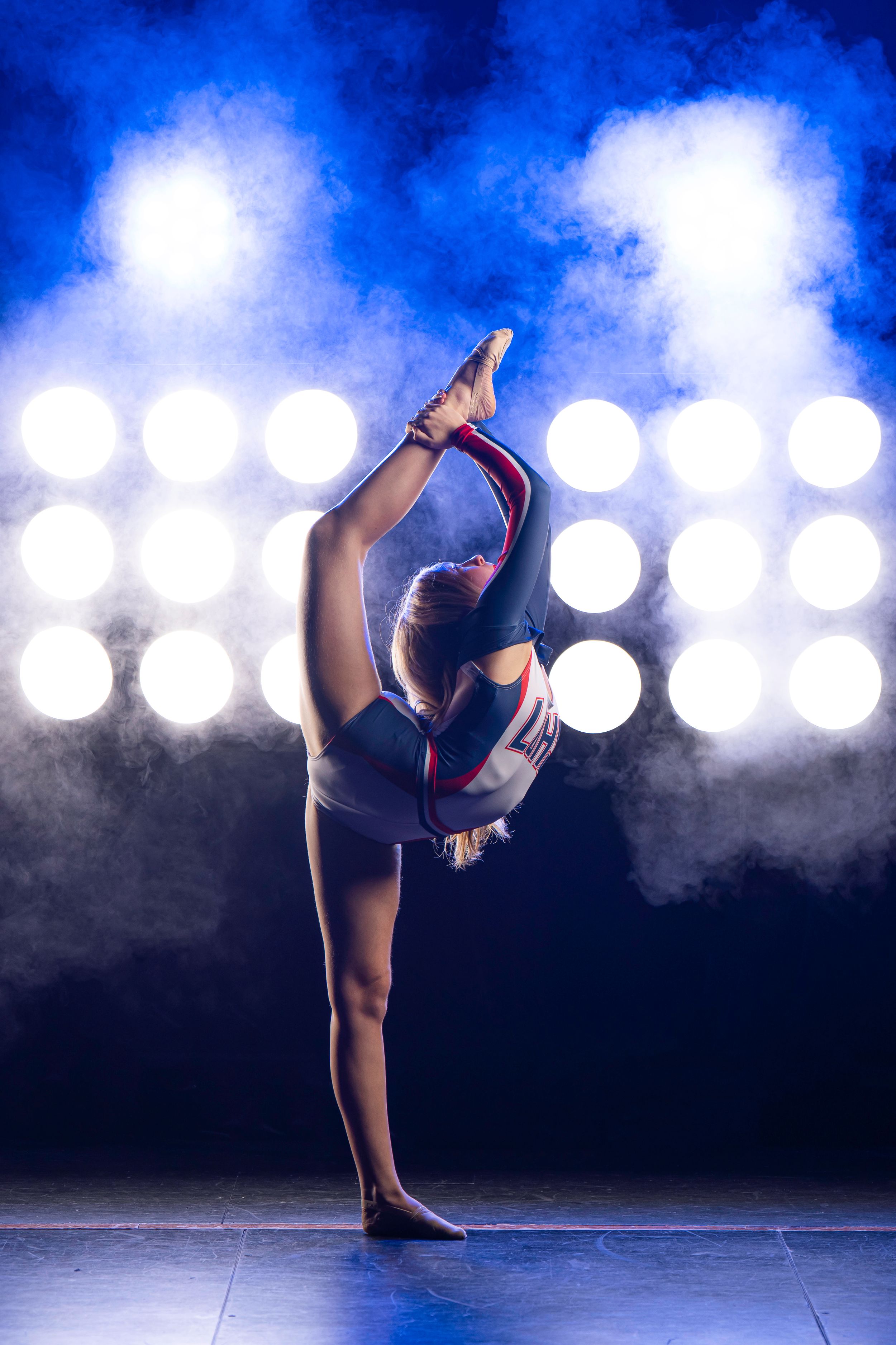 High School Dancer performs graceful split pose on stage with dramatic blue lighting and bokeh spotlight effects in background.