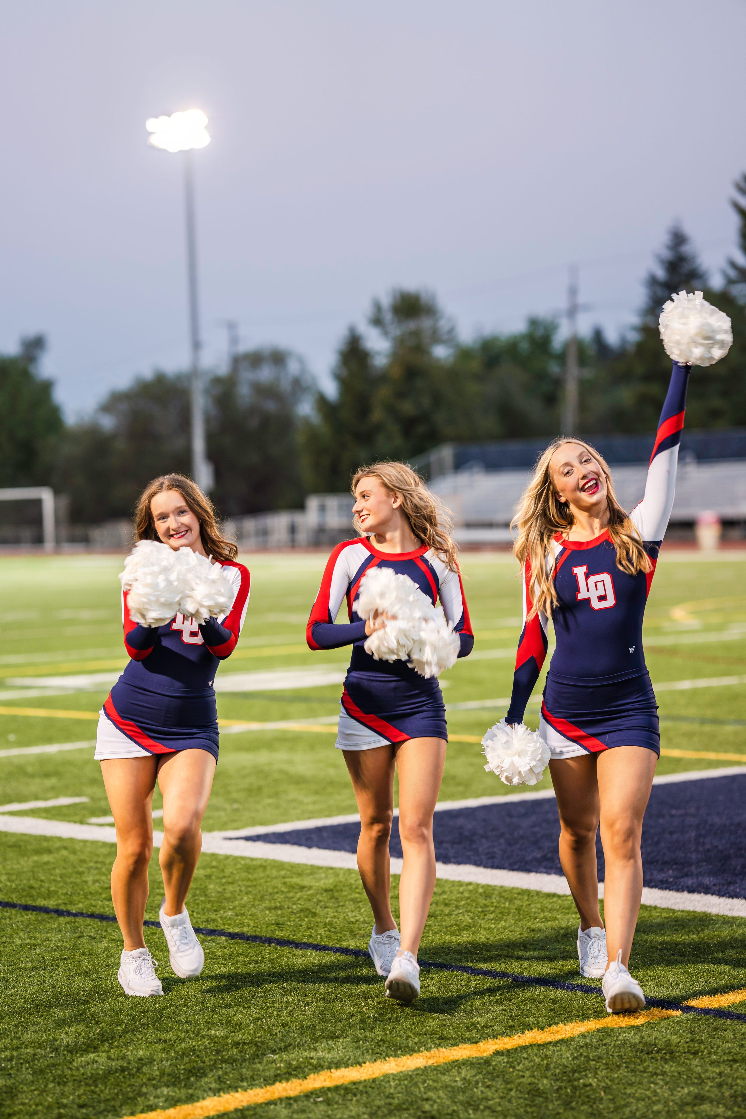 High school senior dance team dancers in navy and red uniforms with pom poms perform on a football field at night under stadium lights.