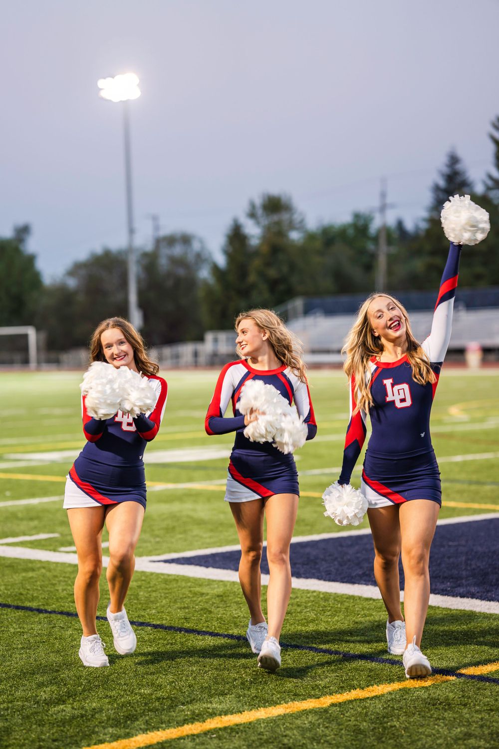 High school senior dance team dancers in navy and red uniforms with pom poms perform on a football field at night under stadium lights.