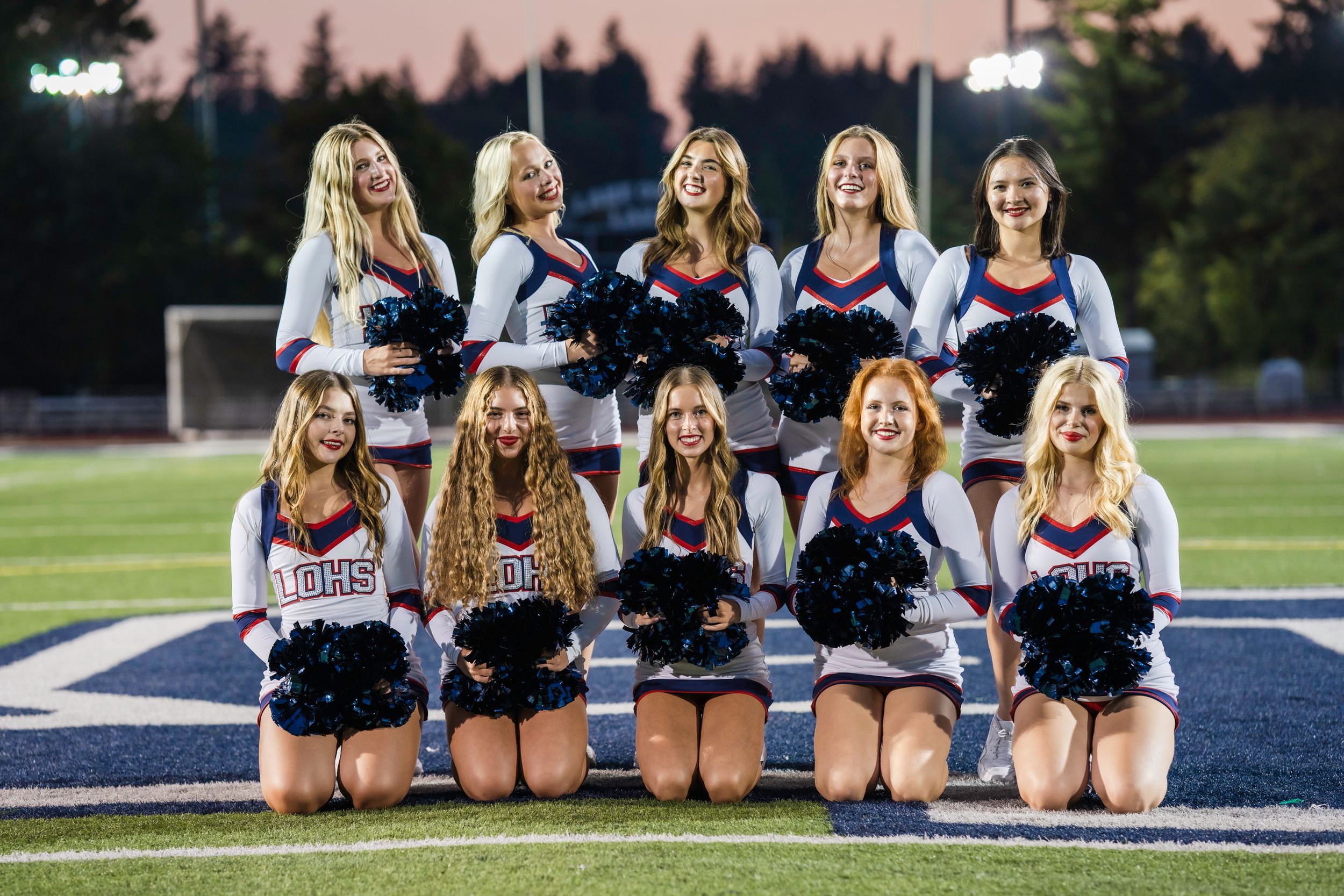 High School dance squad in dark blue and white uniforms poses on football field at dusk with trees in background.