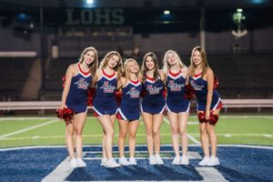 Six high school dance team dancers in navy blue uniforms pose together on a blue football field at night.
