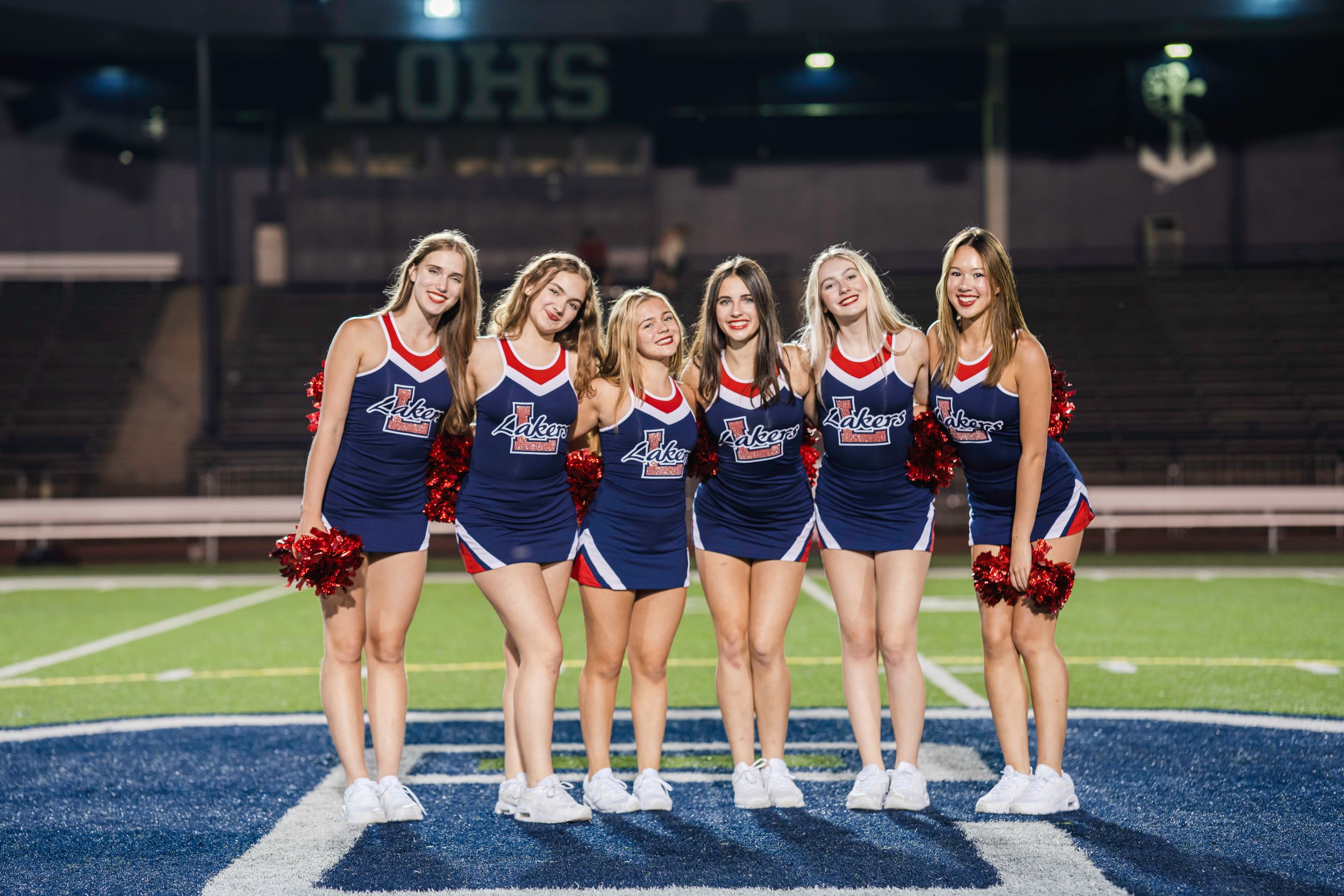 Six high school dance team dancers in navy blue uniforms pose together on a blue football field at night.