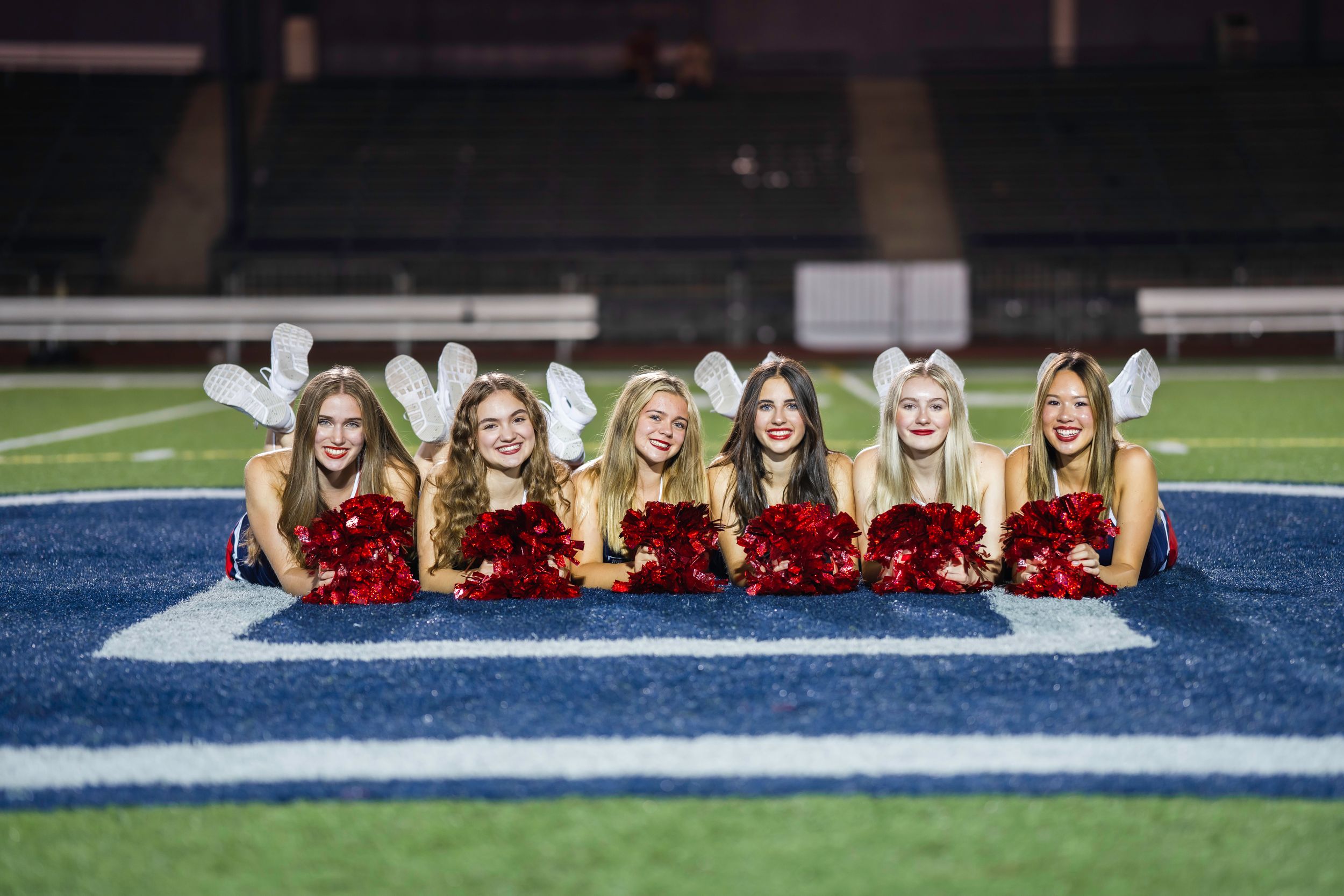High school dancers in uniforms sitting in a line on blue and white striped football field.
