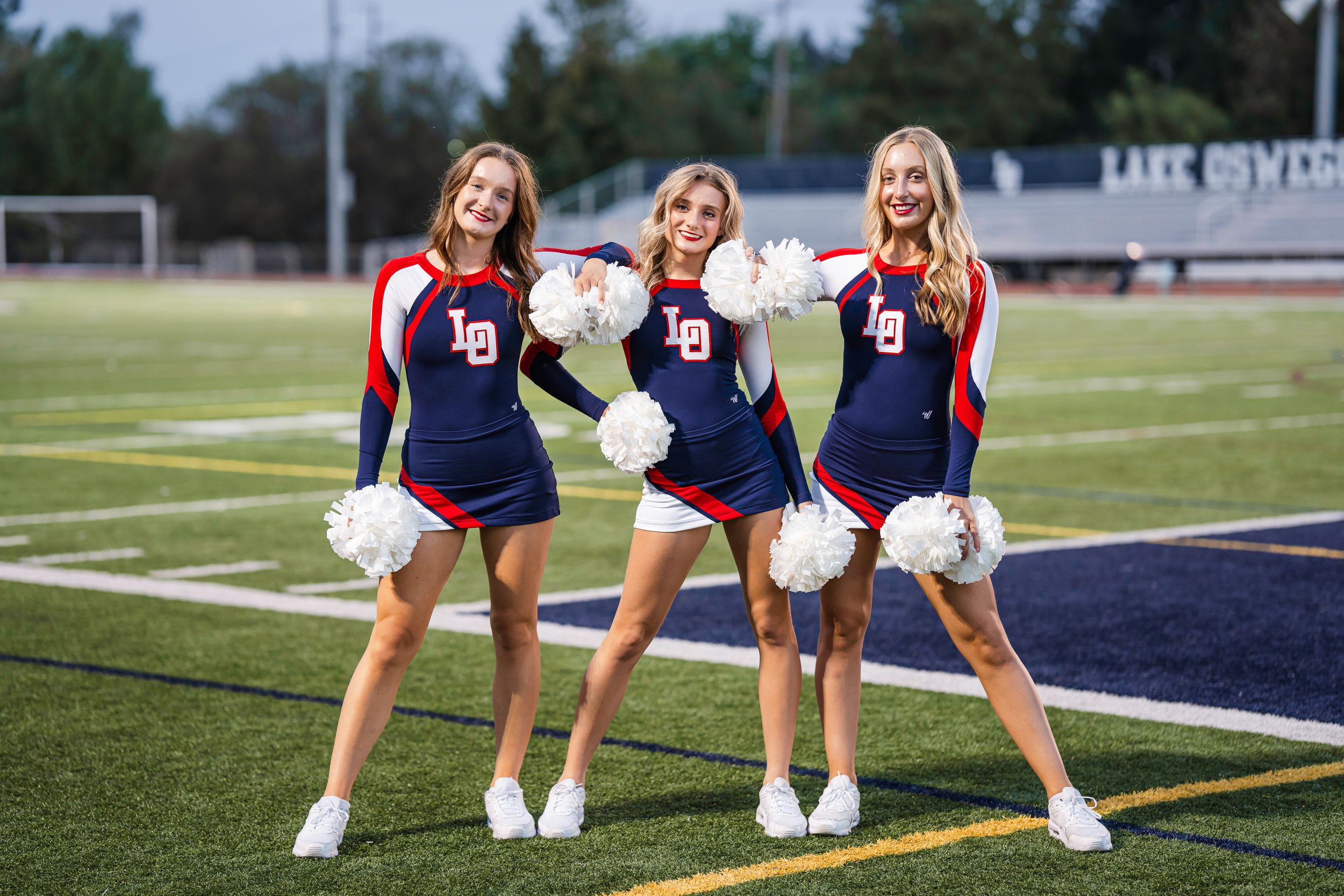 Three cheerleaders in navy uniforms and red capes pose on a sports field with pom poms.