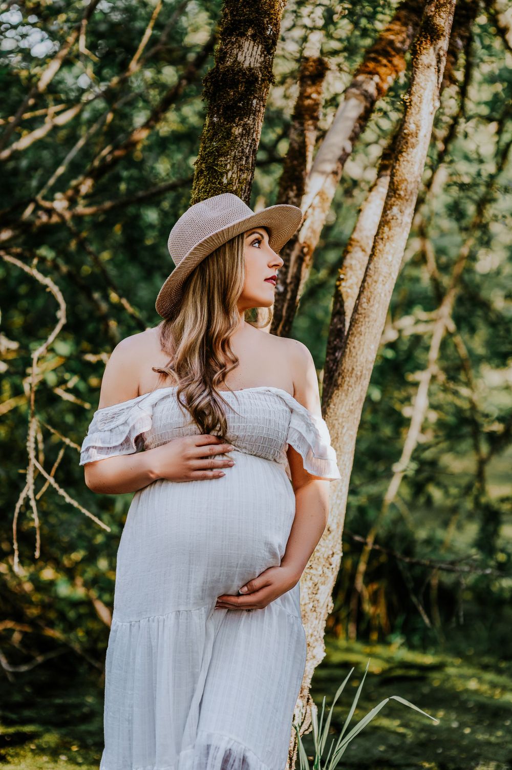 Maternity photo of woman in white dress and hat standing by tree in golden sunlight.