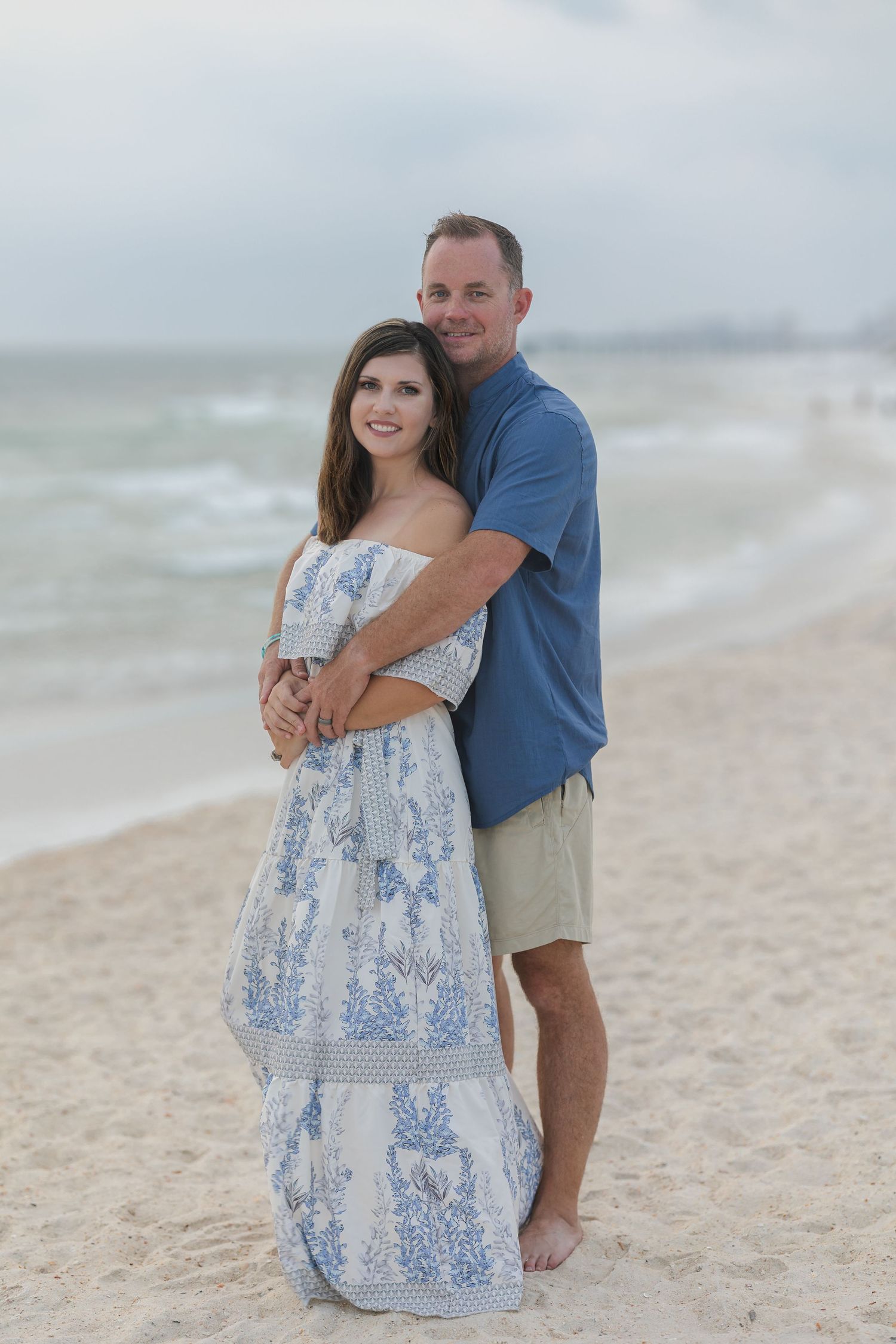 Romantic beach photoshoot featuring coordinated blue and white outfits against ocean backdrop.