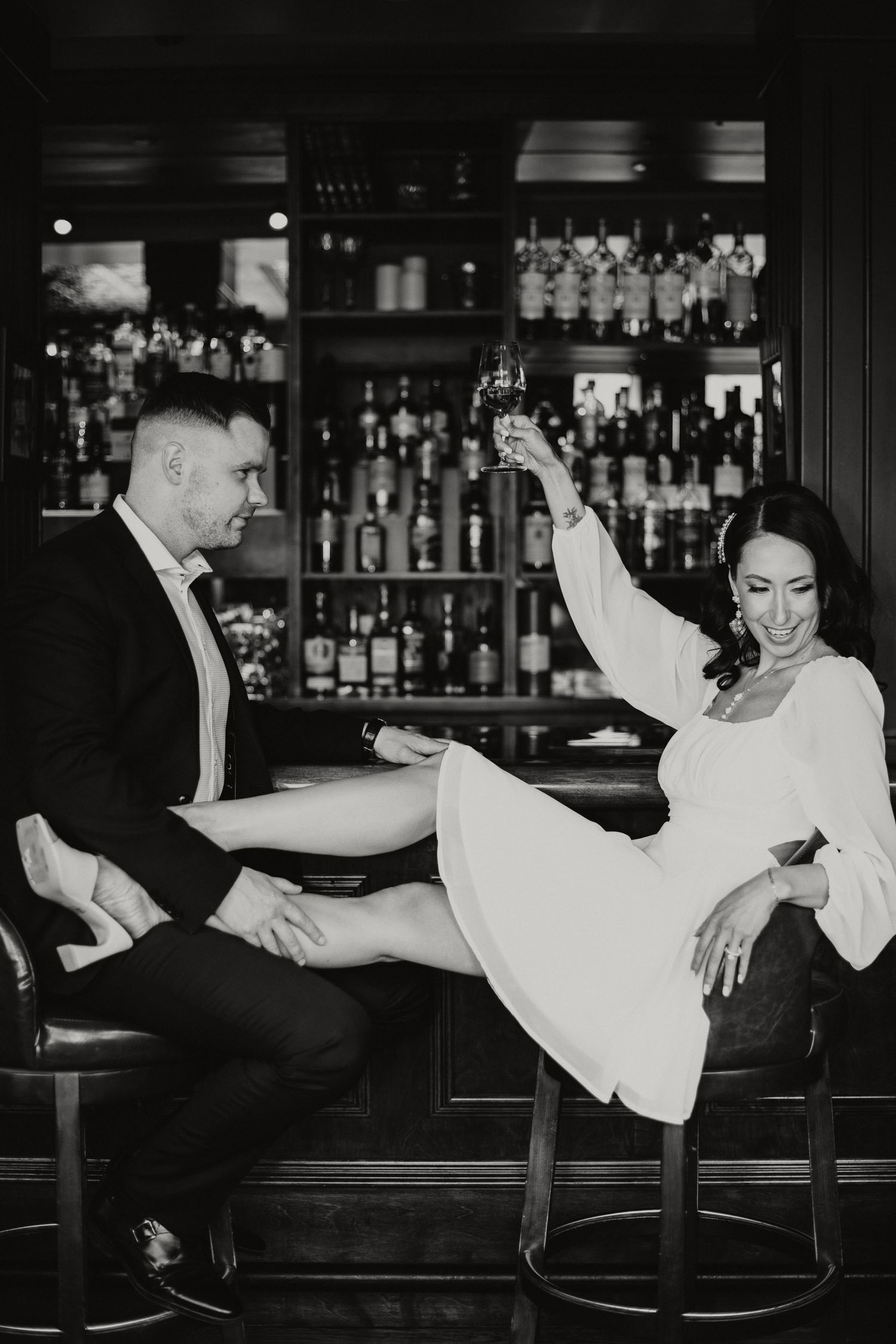 Black and white photo of bride and groom sitting at a bar. The bride is toasting her groom.