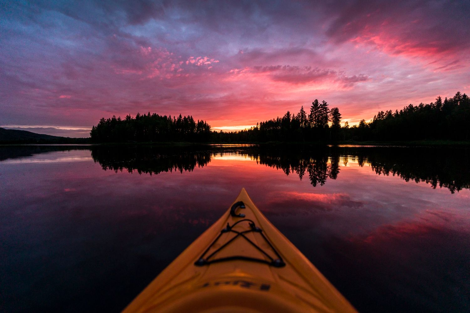 Kayak bow pointing toward a stunning pink and red sunset reflecting on a calm lake.