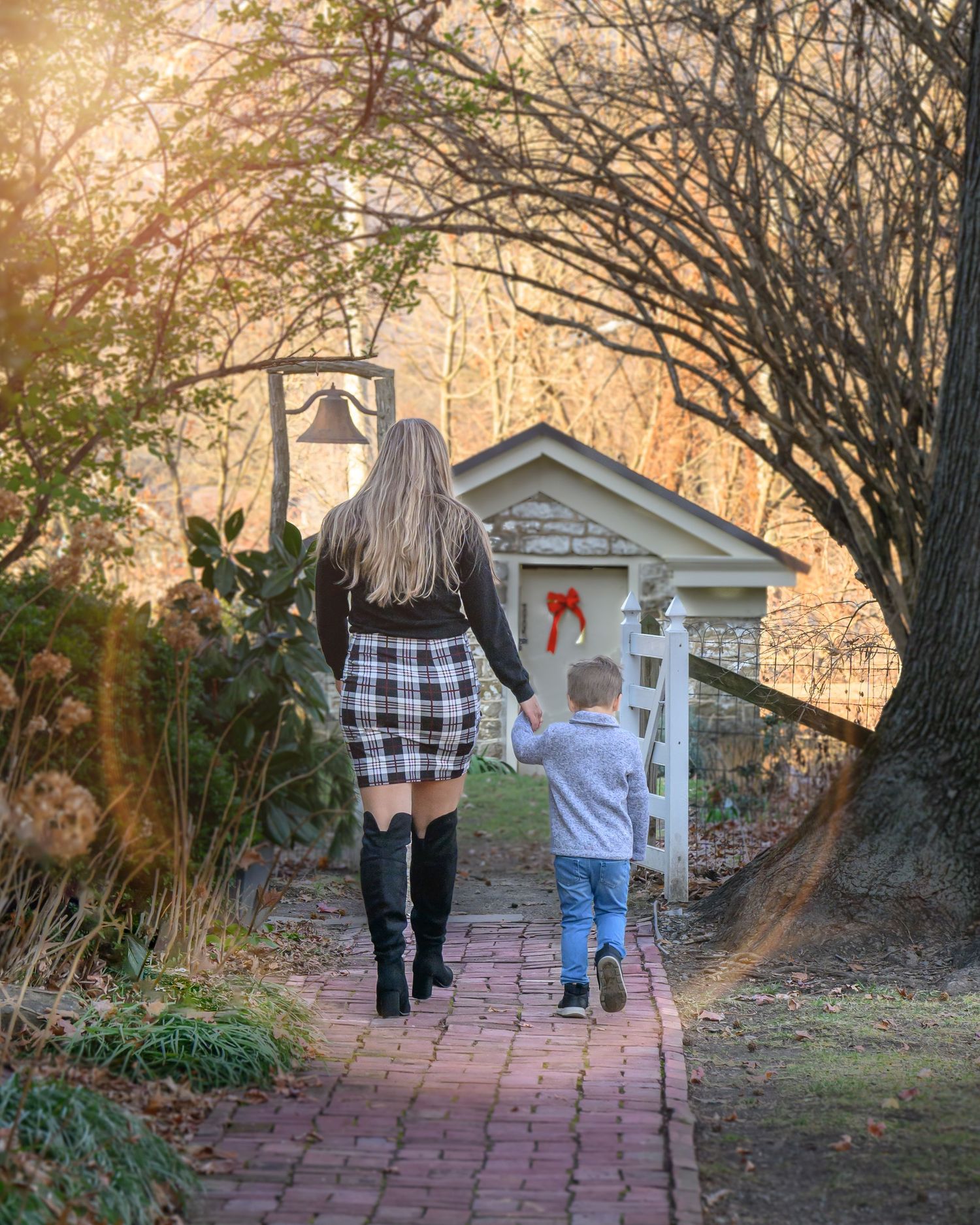 A mother and her son walk down a brick pathway towards a small white garden shed as sunlight filters through tree branches.