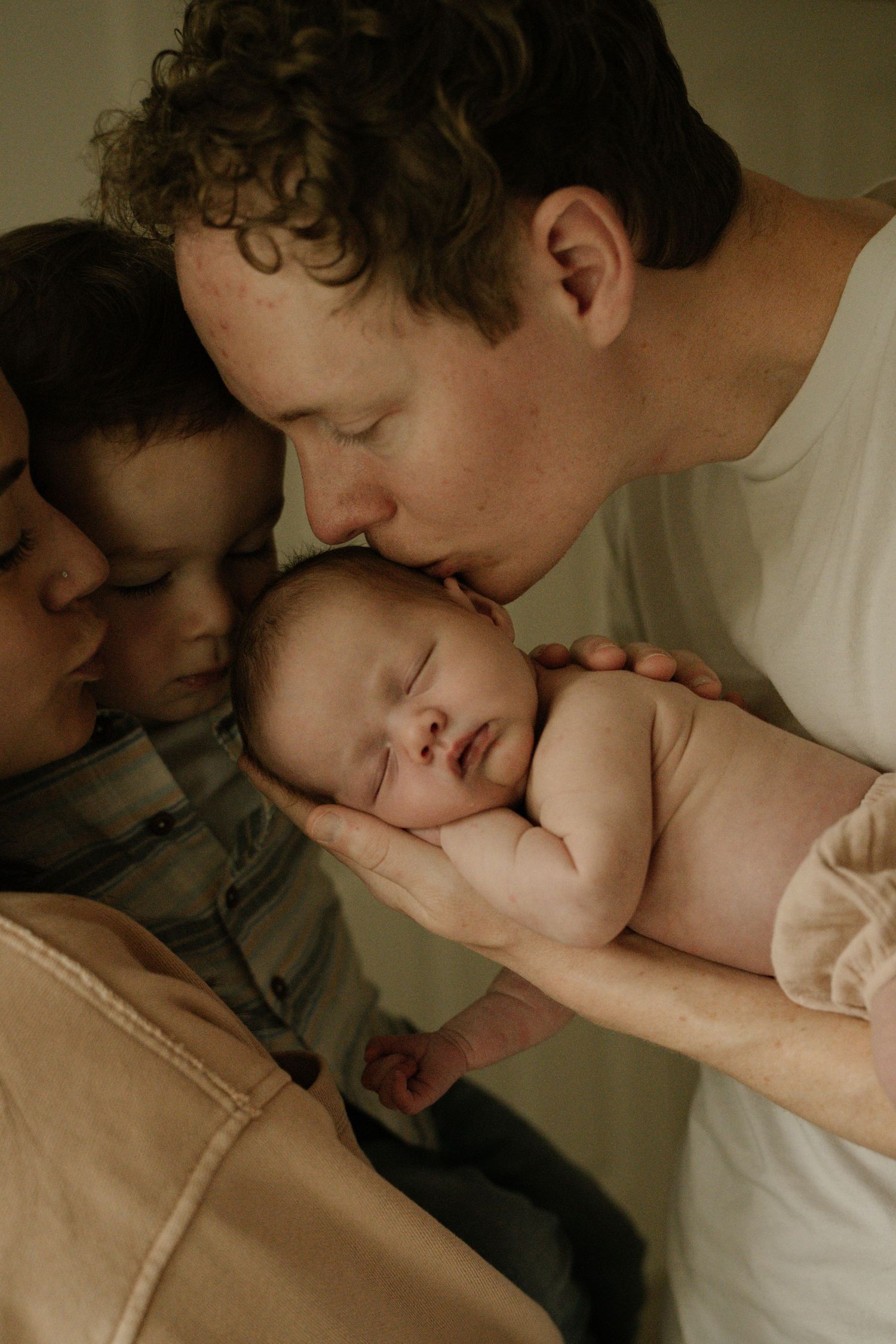 Father gently kisses sleeping newborn baby held in hands while family gathers closely together indoors. Victoria, BC, Vancouver Island.