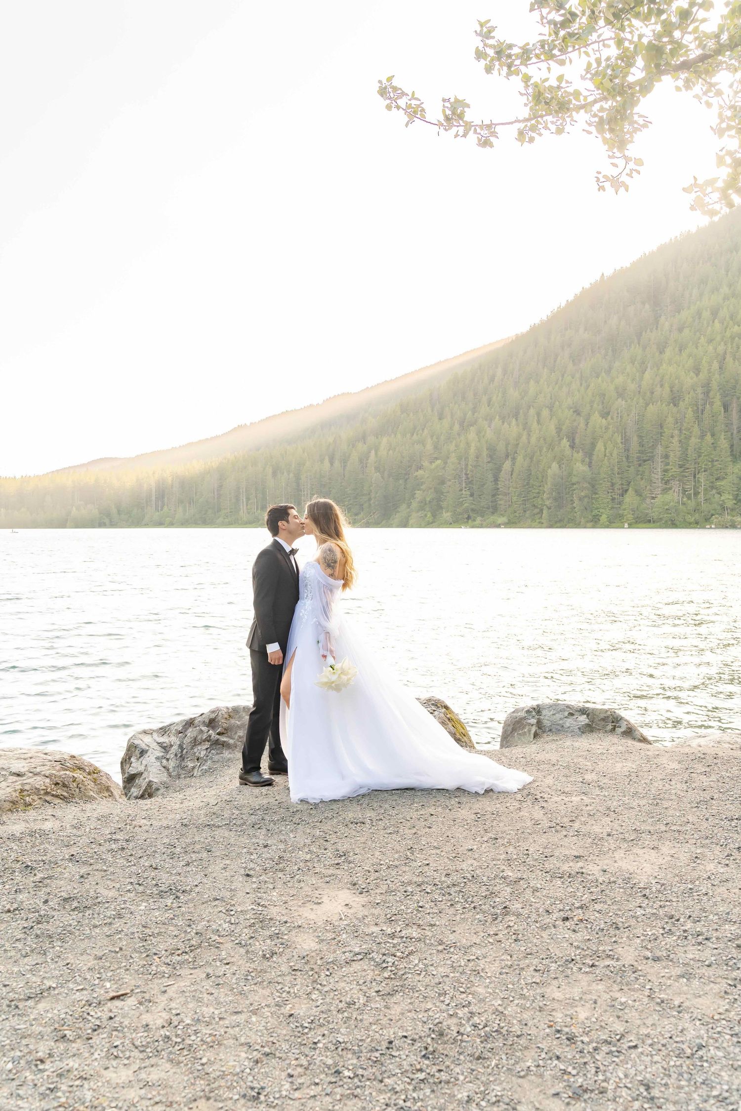 A romantic wedding couple embraces by a serene mountain lake at sunset with evergreen forests in the background.