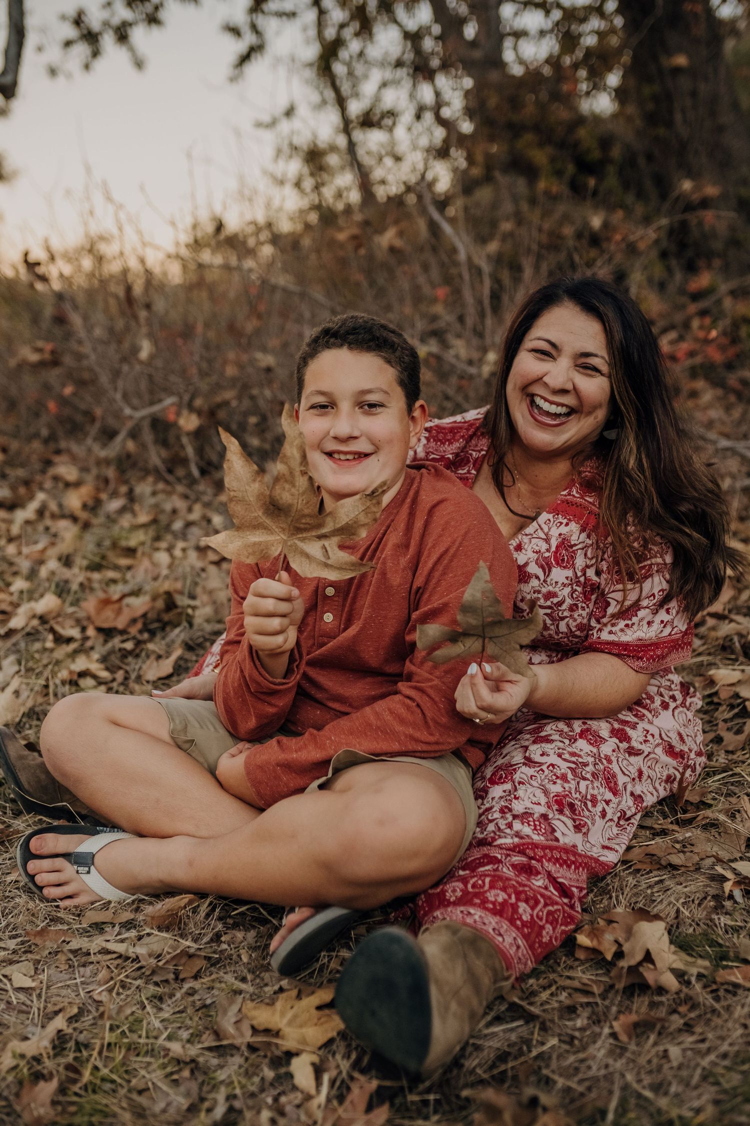 A mom and her son sit on the ground together while both holding large maple leaves.