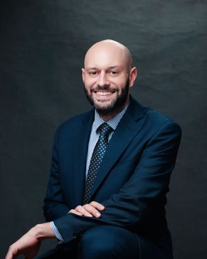 Professional headshot of a person in a dark blue suit and polka dot tie seated against a gray background.