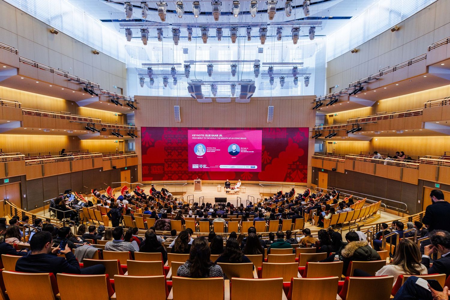 A large modern concert hall filled with audience members facing a red illuminated stage during a formal conference event.