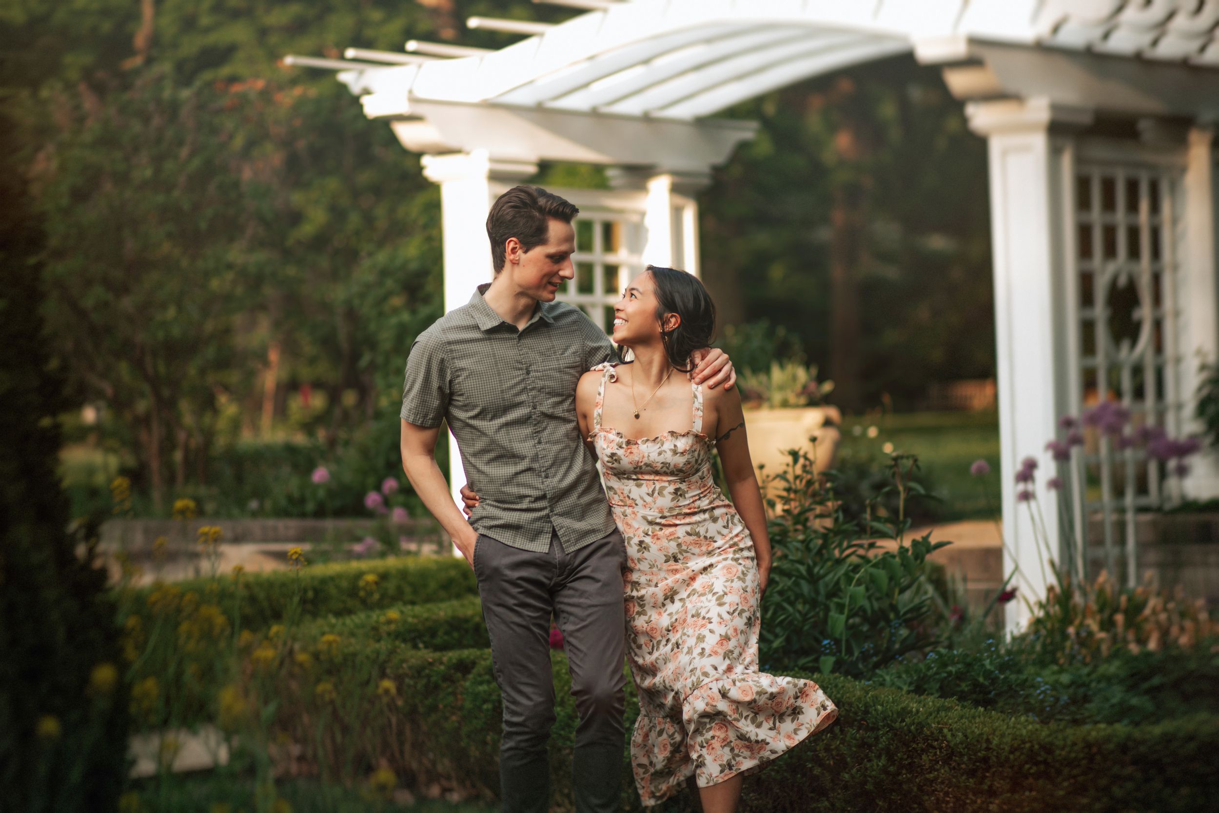 A couple shares a romantic moment in an outdoor garden setting with white pillars and lush greenery in the background.