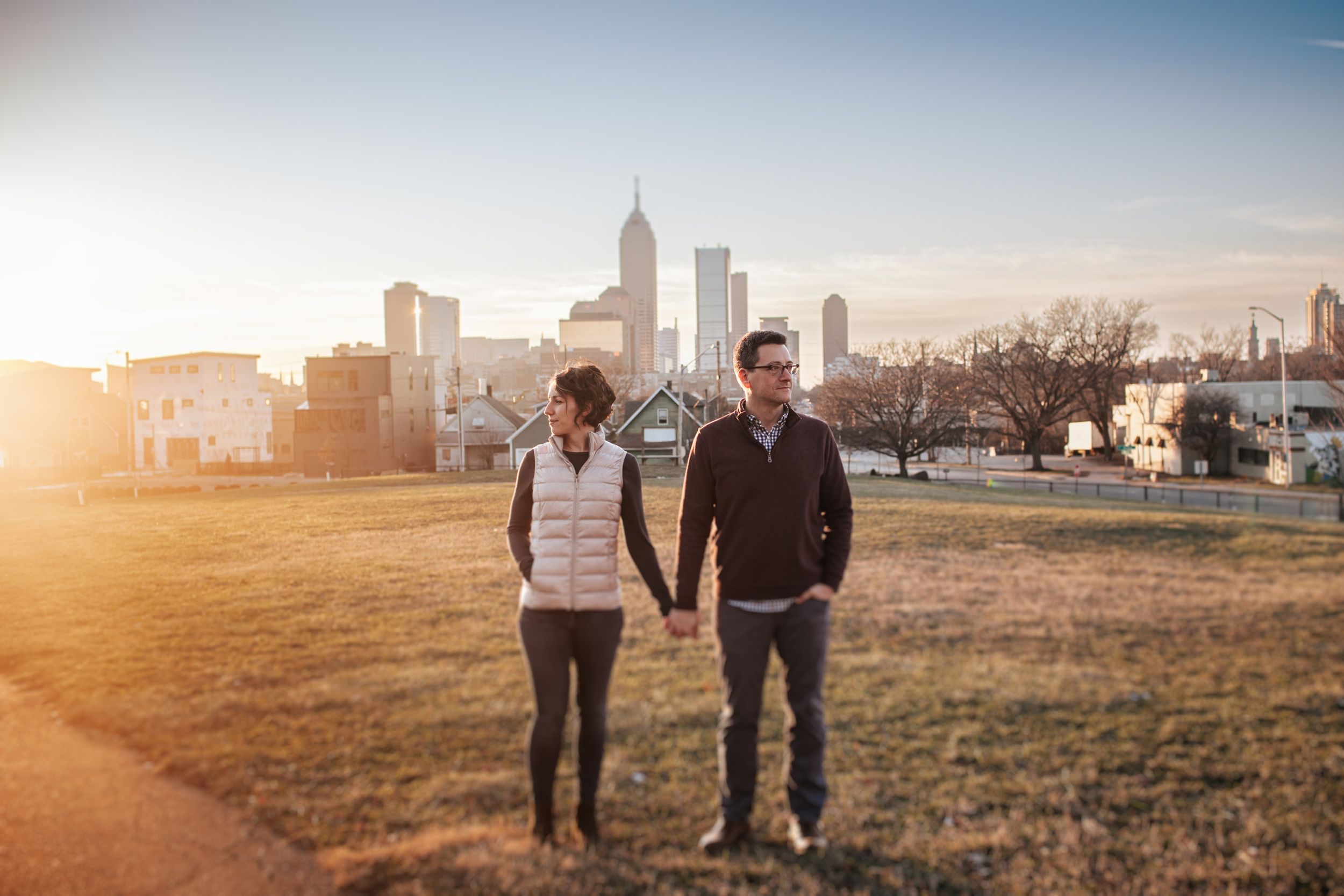 Couple walking together on grassy field at sunset with city skyline in background.