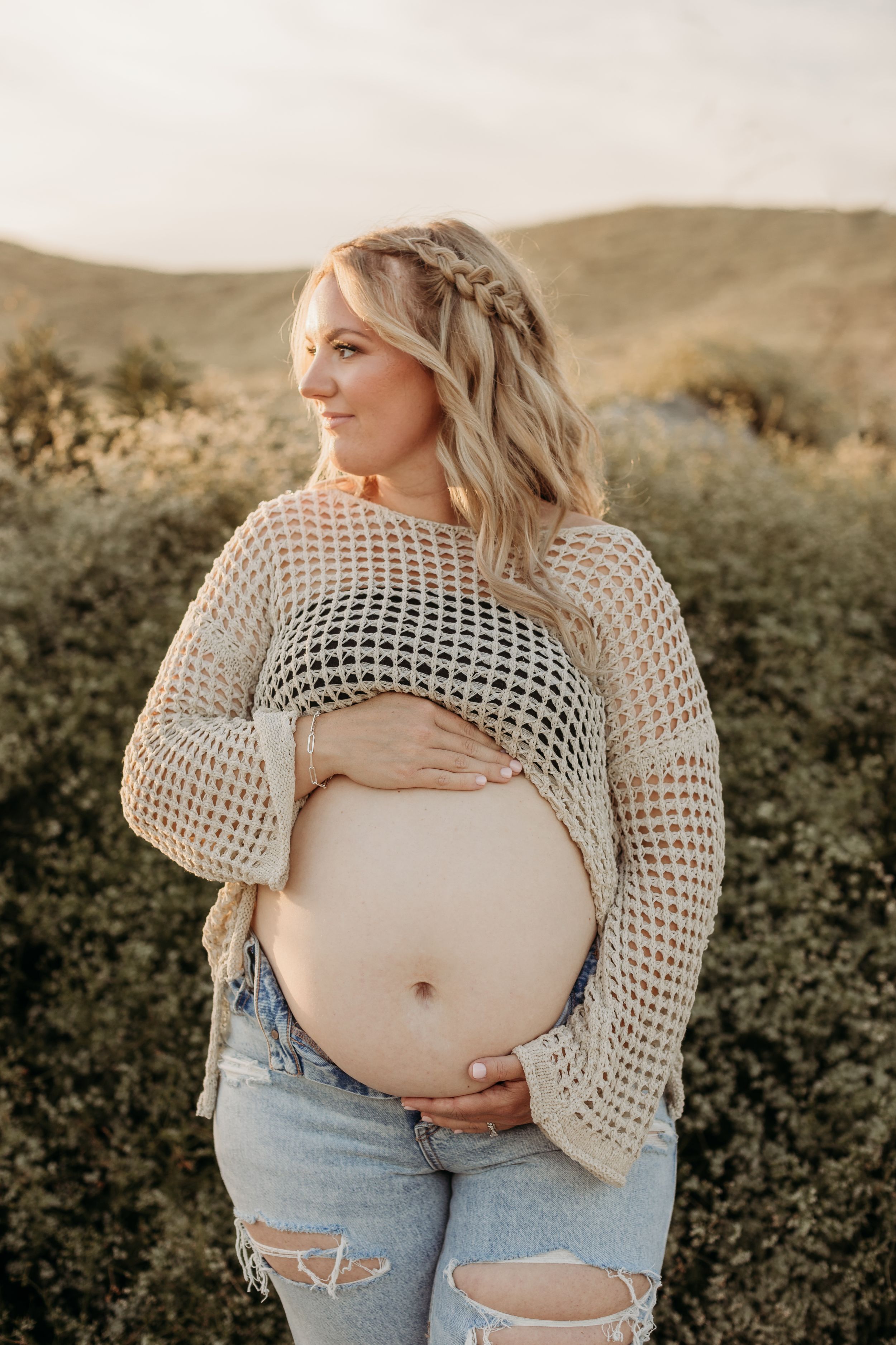 A maternity photo showing a woman in a crochet top and distressed jeans against nature.