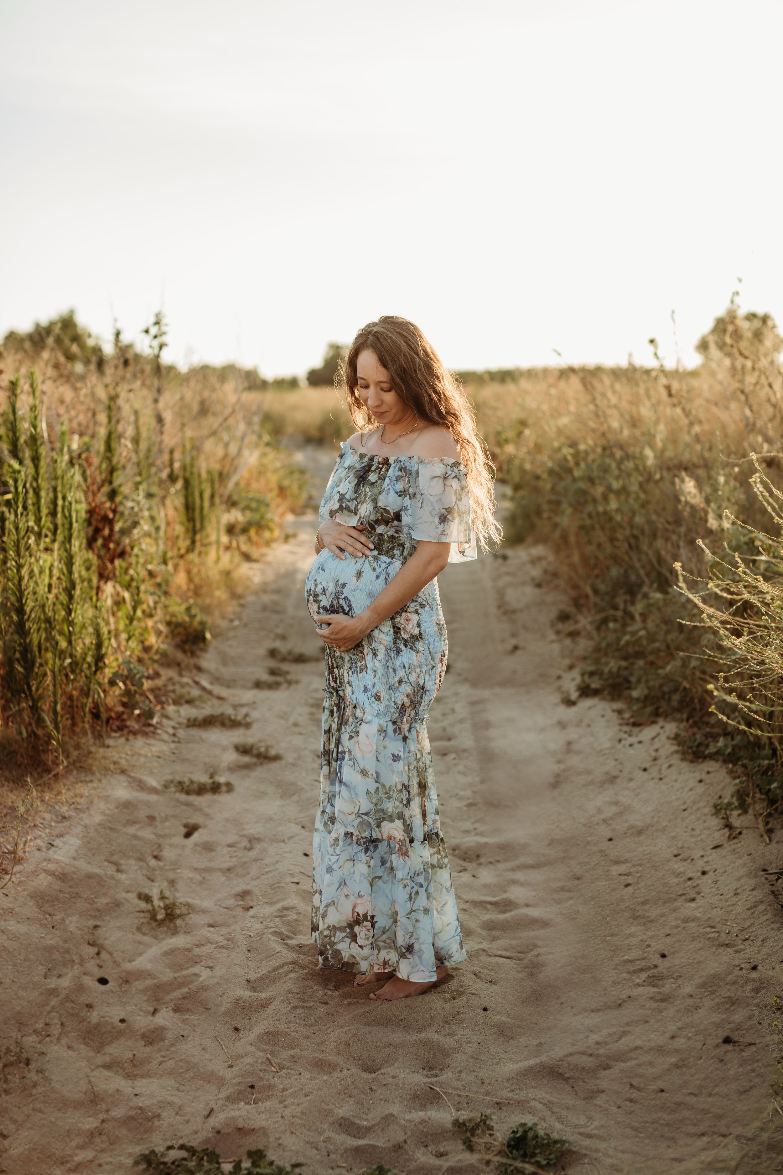 A pregnant woman in a floral maxi dress poses on a dirt path during golden hour.