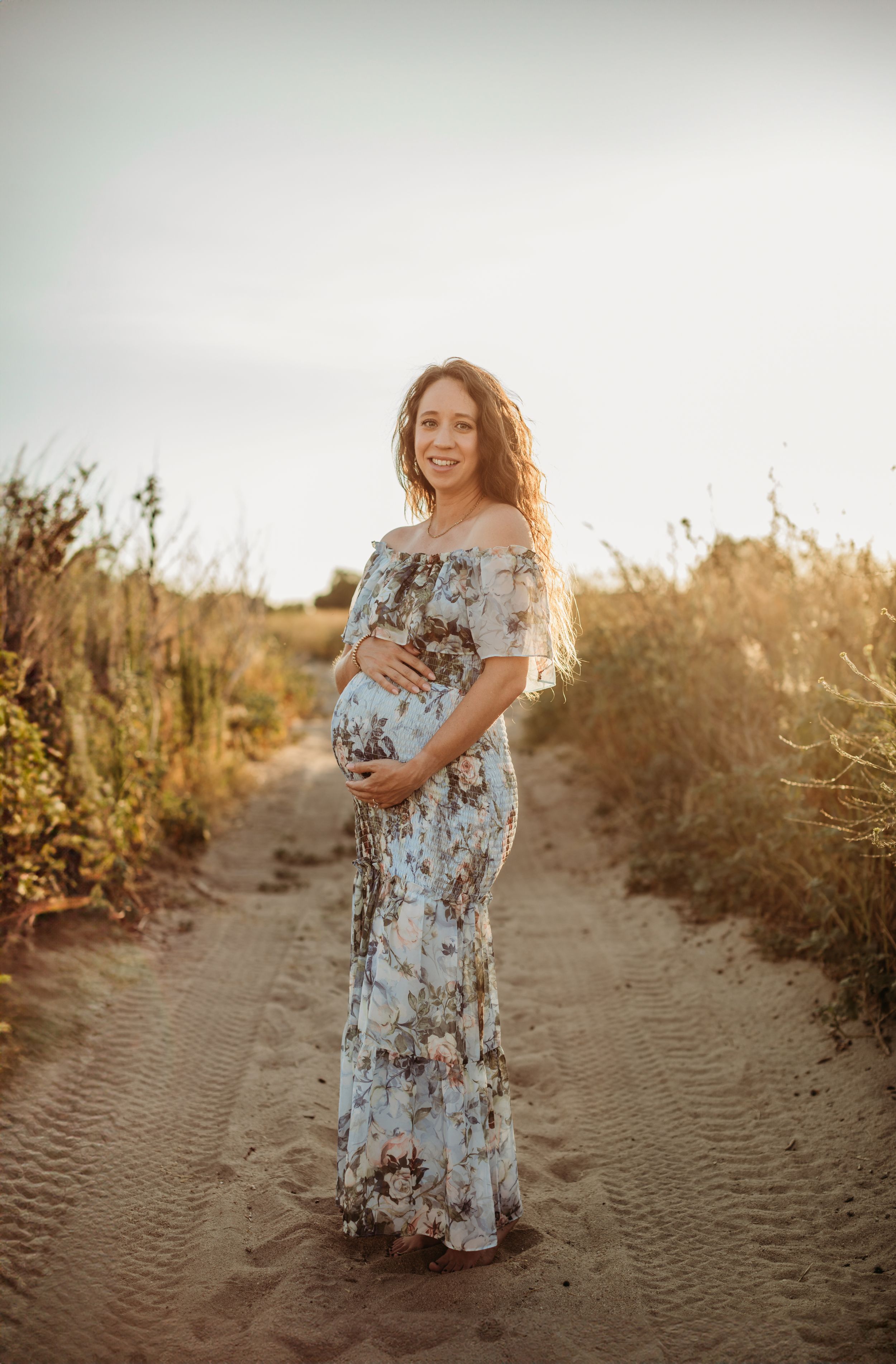 An expectant mother in a floral dress poses during sunset on a sandy trail.