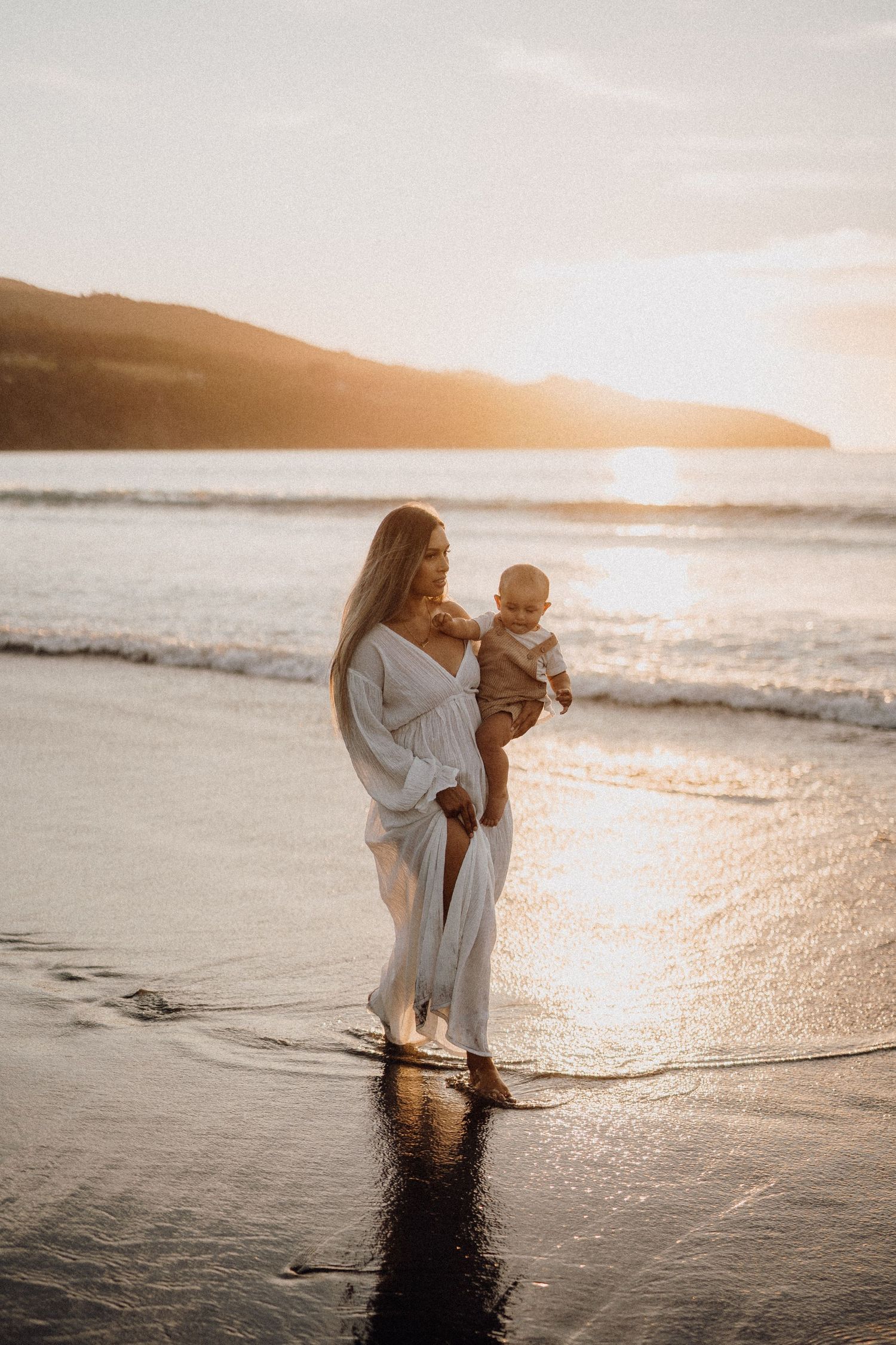 An expectant parent in a flowing dress stands on a beach at sunset.
