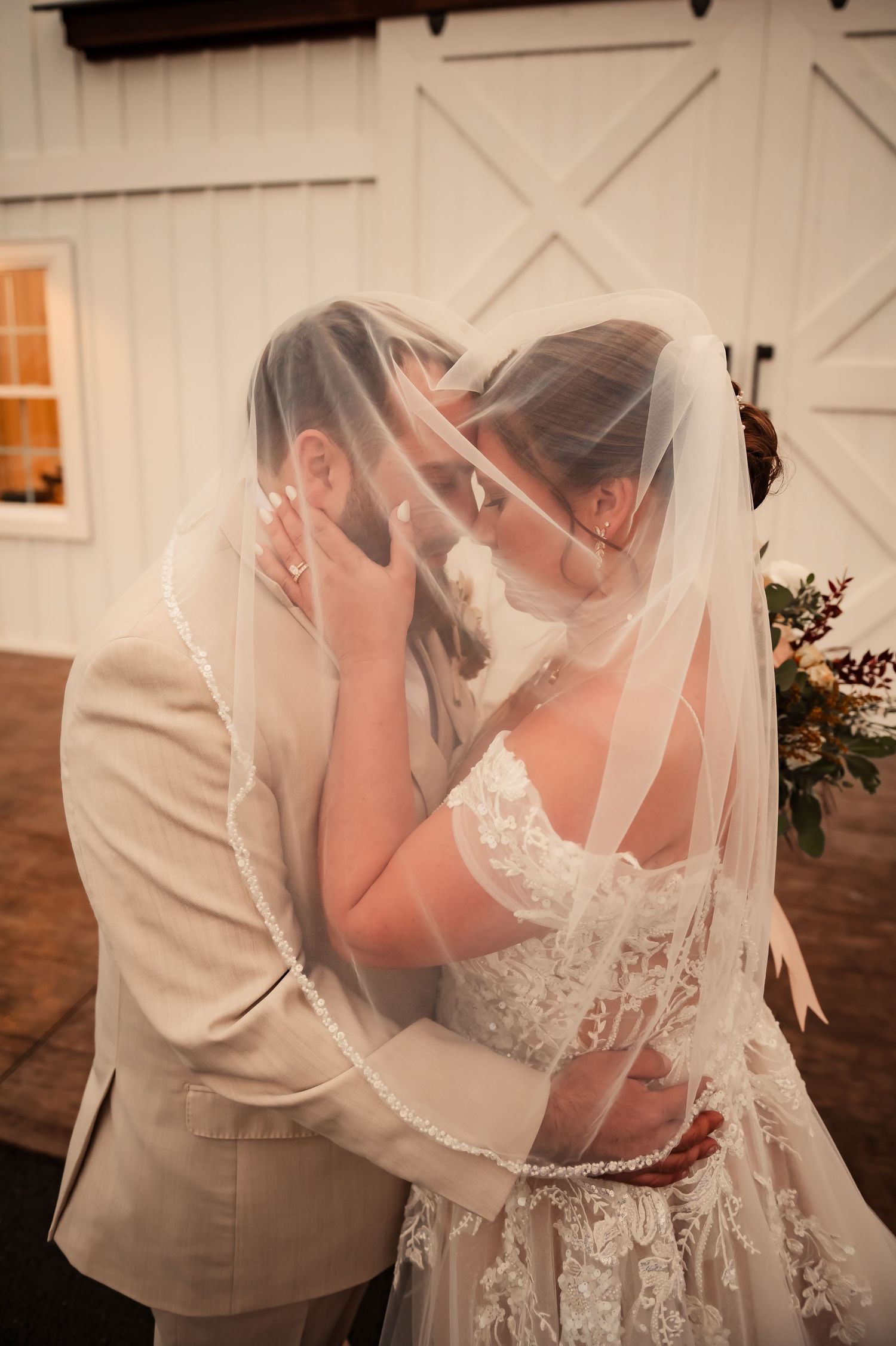 Romantic wedding moment with couple embracing under delicate lace veil against barn backdrop.