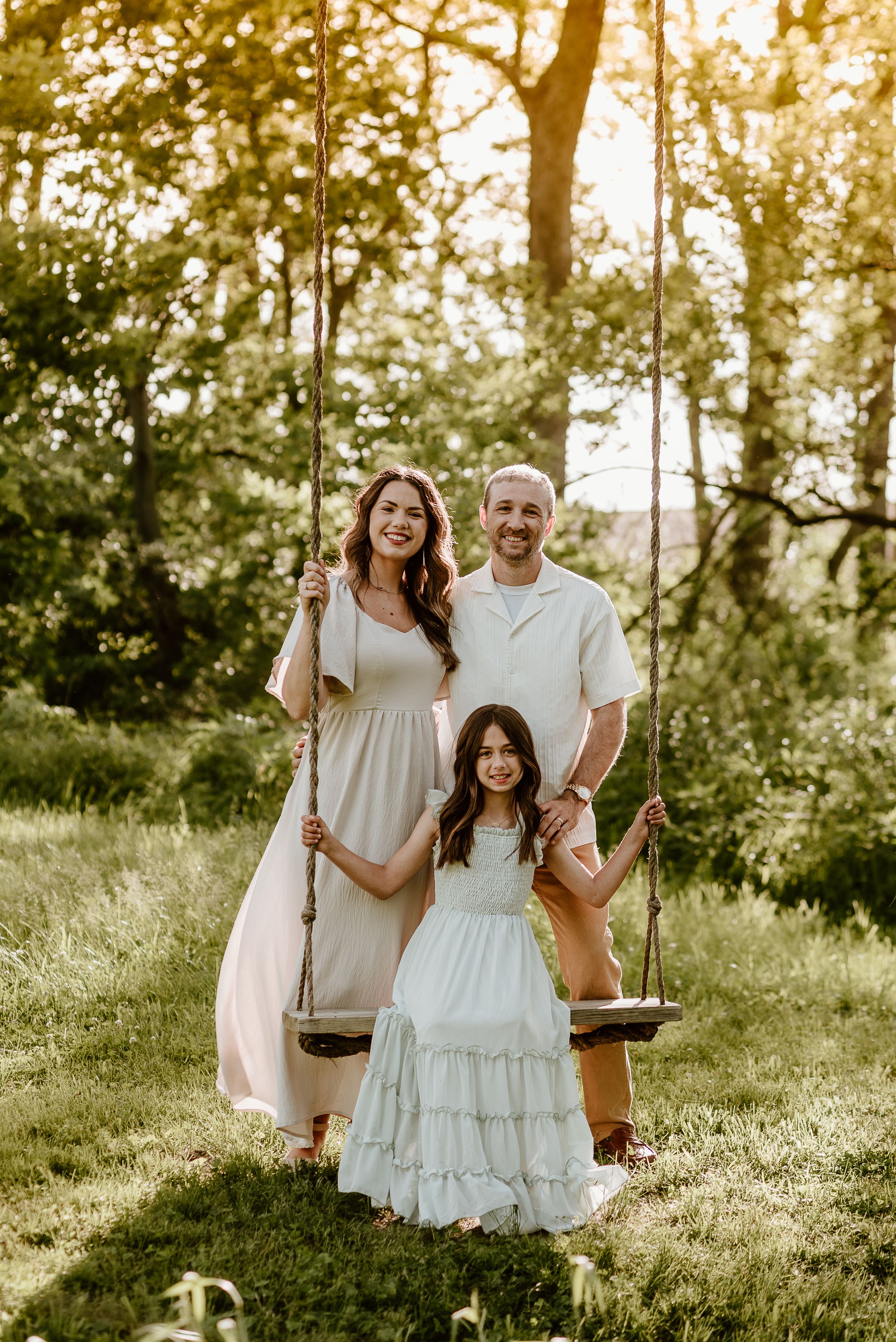 little girl on a wooden tree swing, her parents standing behind her
