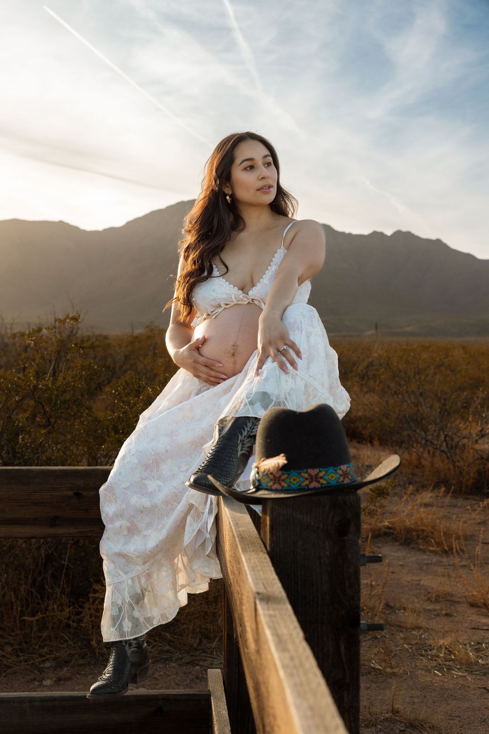 Person in white dress poses on wooden fence at sunset in desert landscape with mountains in background.