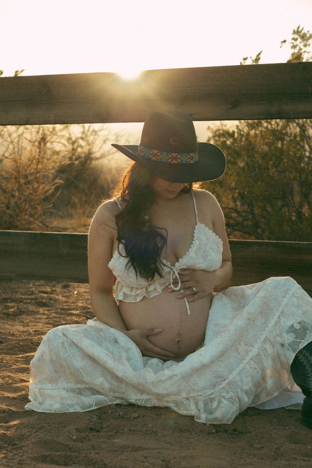 A couple in western attire shares a romantic moment at sunset while sitting on the ground in a desert landscape.