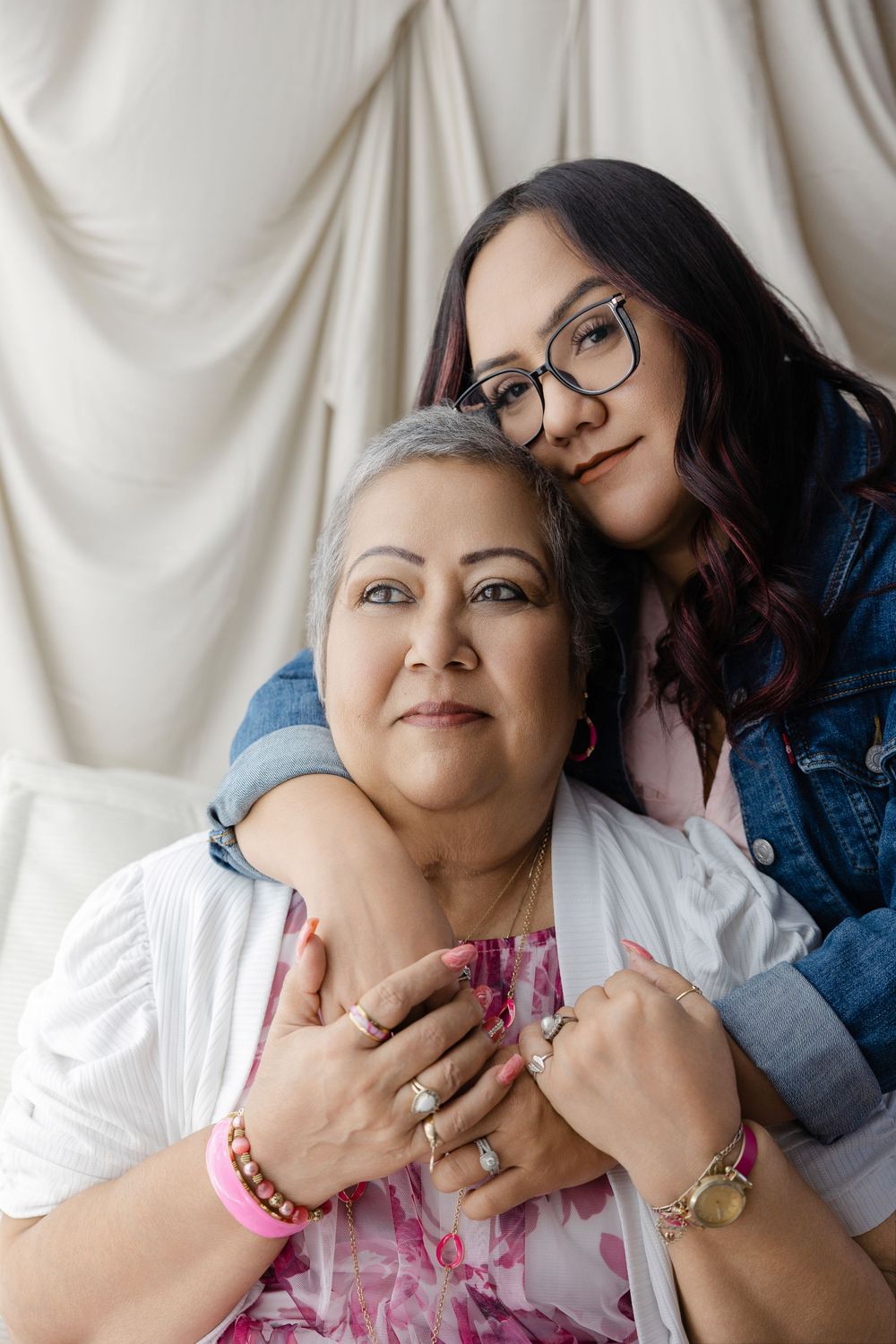 Two women embrace warmly in a studio portrait, younger woman with glasses hugging older gray-haired woman from behind.
