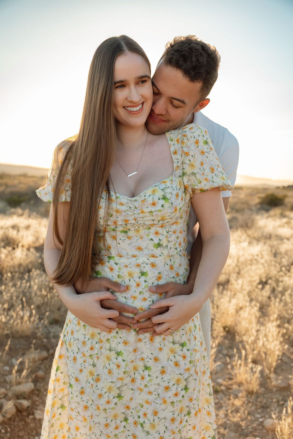 A couple embraces in a sunny field, with her wearing a yellow floral dress during a romantic sunset photoshoot.