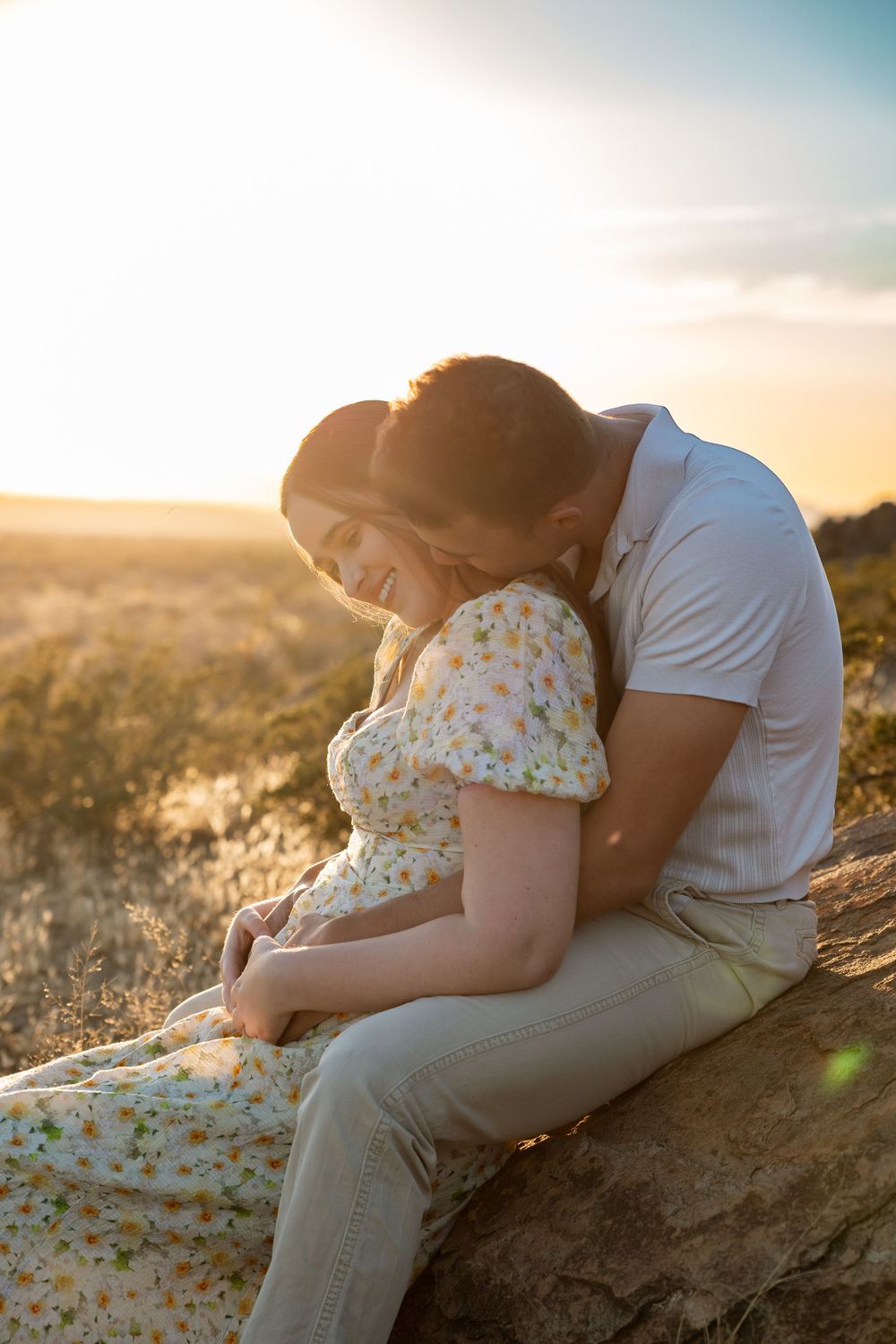 Romantic couple embraces on a hillside at golden hour as sunlight filters through tall grass in the background.