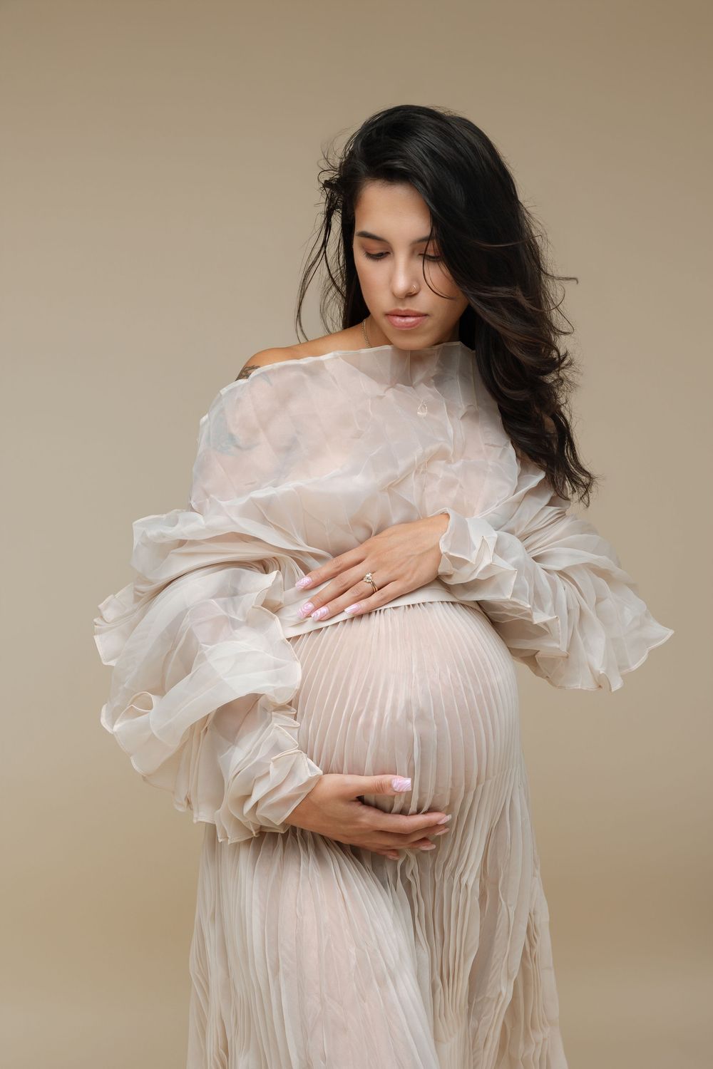 Pregnant woman in sheer beige off-shoulder gown cradling baby bump, dark hair, studio portrait on neutral background.