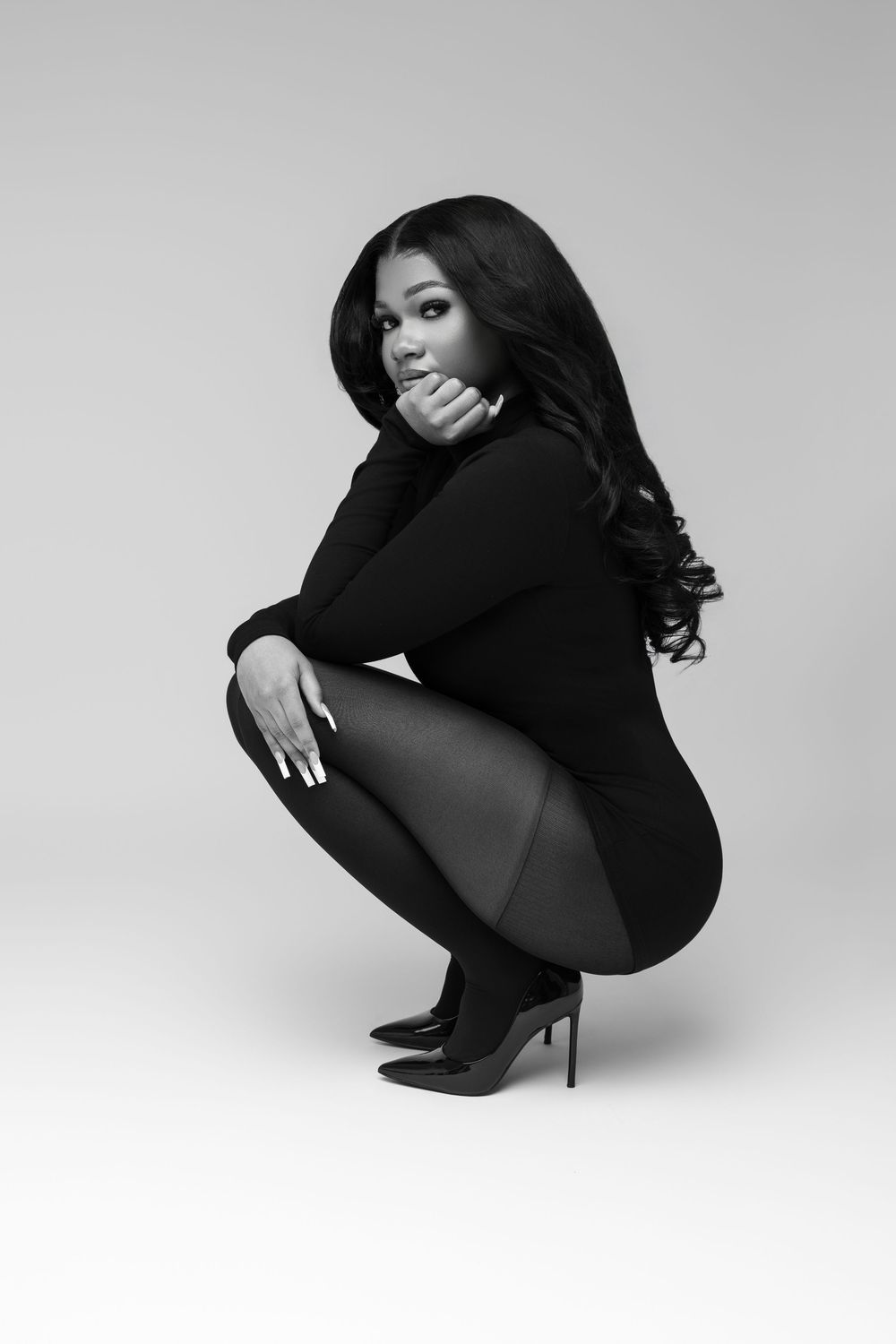 Woman in black outfit and heels crouching gracefully in a sleek black and white studio portrait.