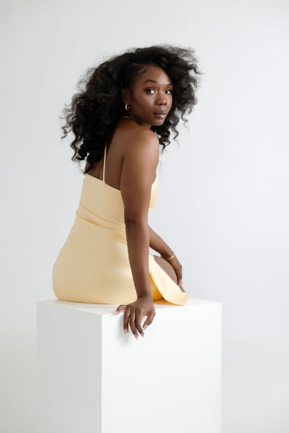Young Black woman in yellow mini dress sitting on white cube, curly hair flowing, studio photo.