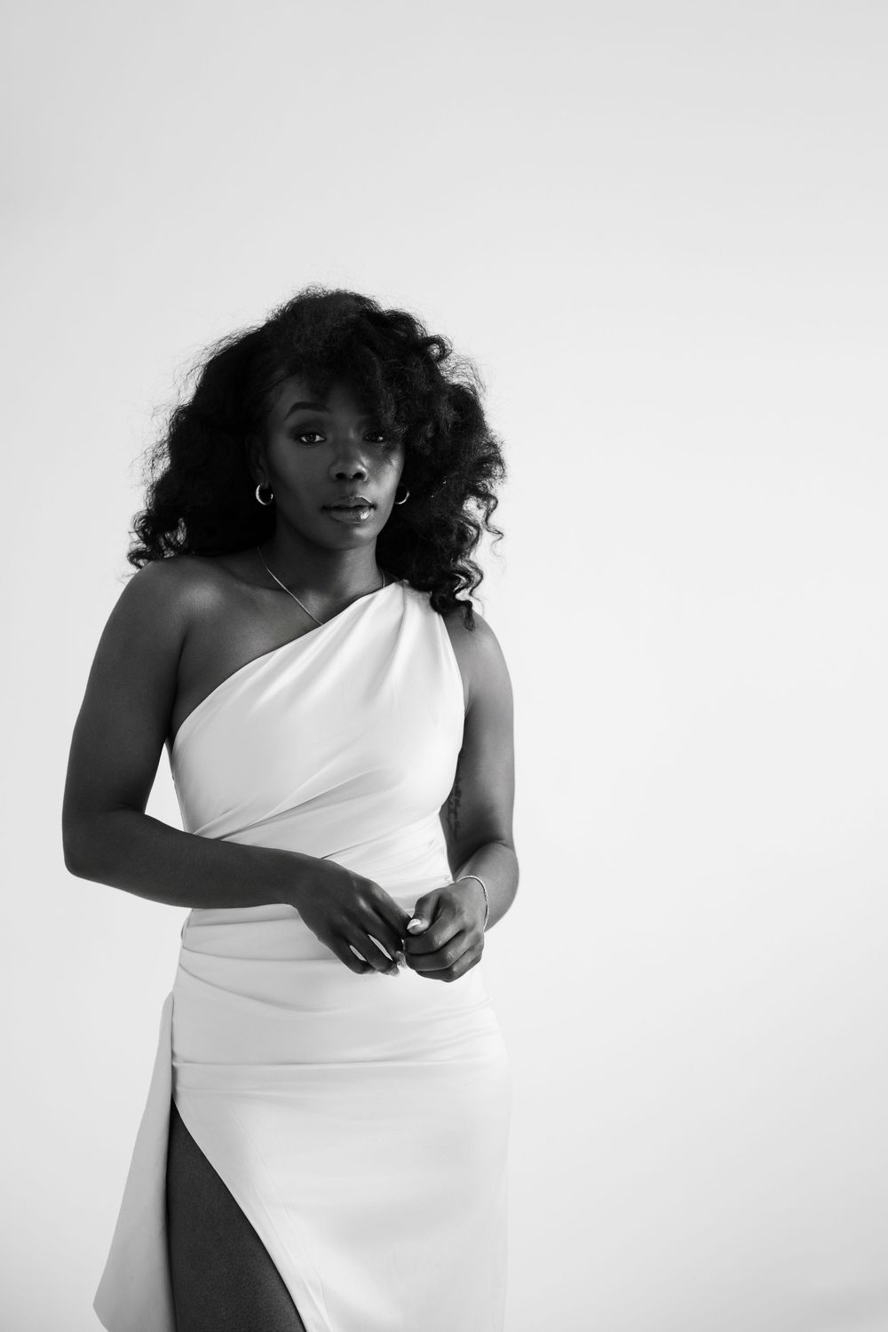 Black and white portrait of a woman in an elegant one-shoulder white dress, posing confidently against a light background.