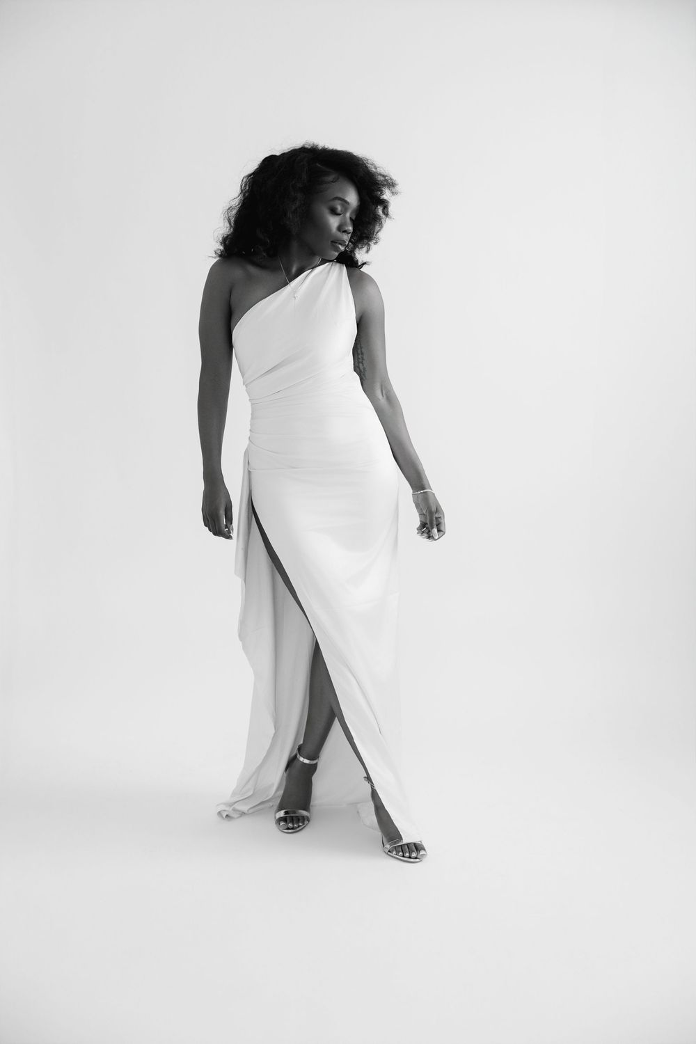 Black and white photo of a woman in an elegant one-shoulder white gown with a slit, posing gracefully in heels.