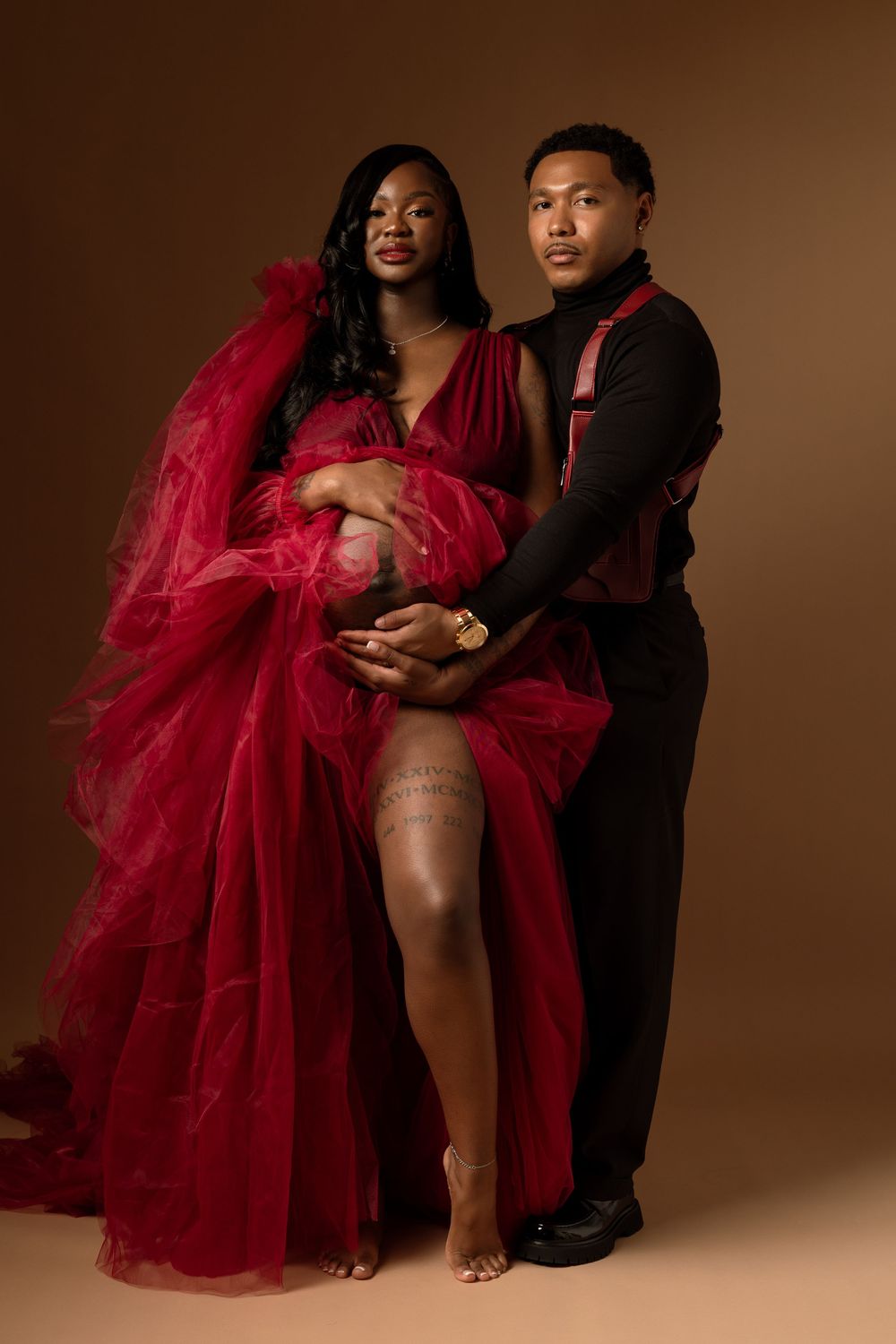 Professional maternity photoshoot featuring couple in black and red attire against warm brown backdrop.