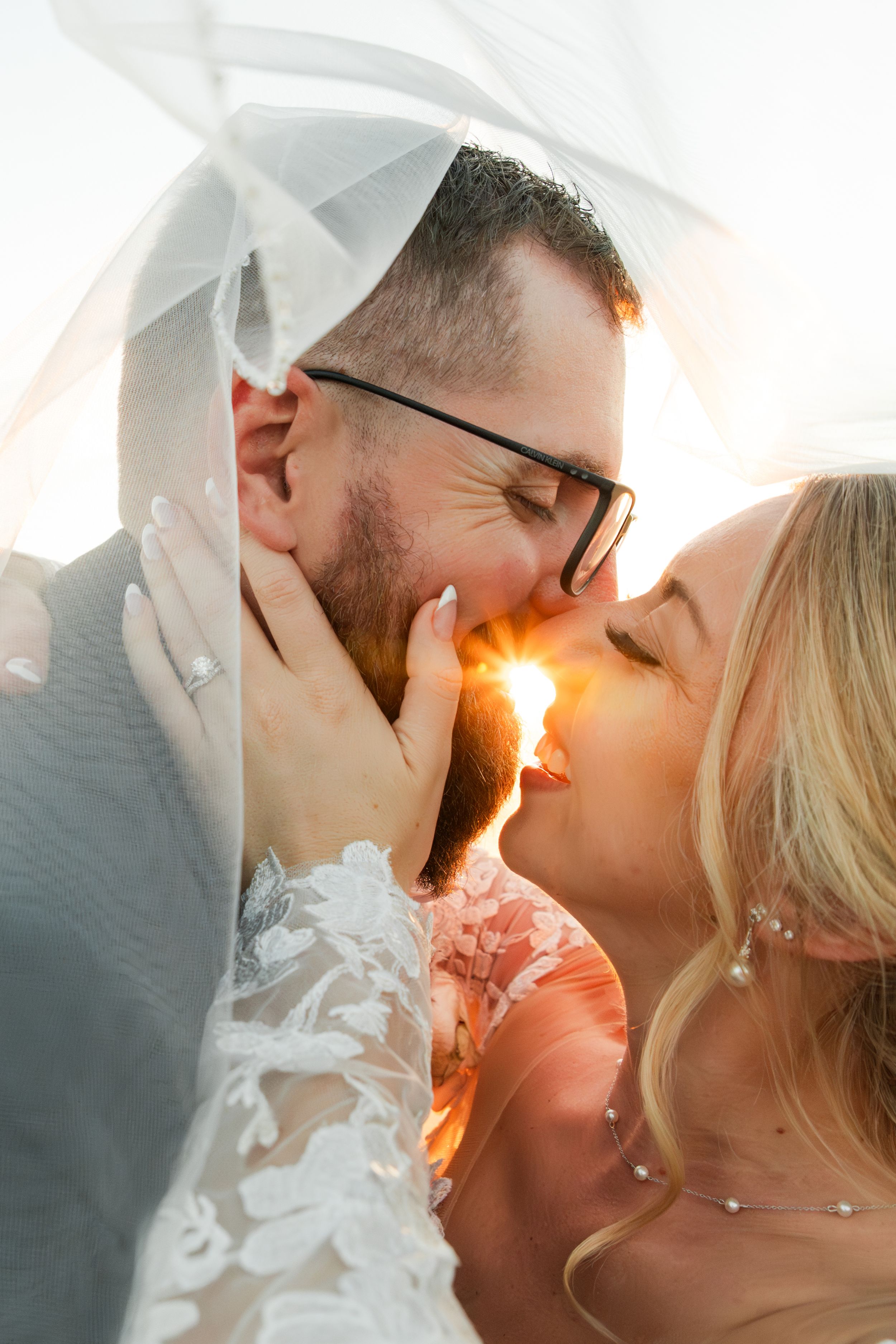 A romantic wedding photo with a couple kissing in golden sunlight through flowing white veil.